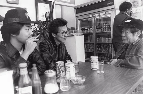 Black and white screenshot of three Chinese men sitting around a table at a cafe with some cans and glasses in front of them.
