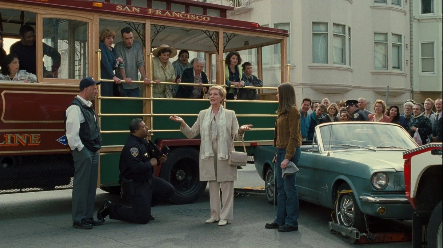 Screenshot from The Princess Diaries, Julie Andrews speaks to a group of people in front of a cable car as a policeman kneels in front of her.
