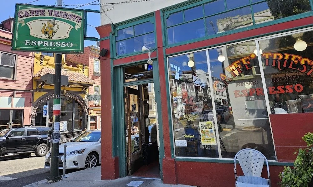 A dark red and green building on a street corner with the door open. A sign on a pole outside the building reads "Caffe Trieste Espresso" with an old coffee pot detailed on it. The door to the building is open.