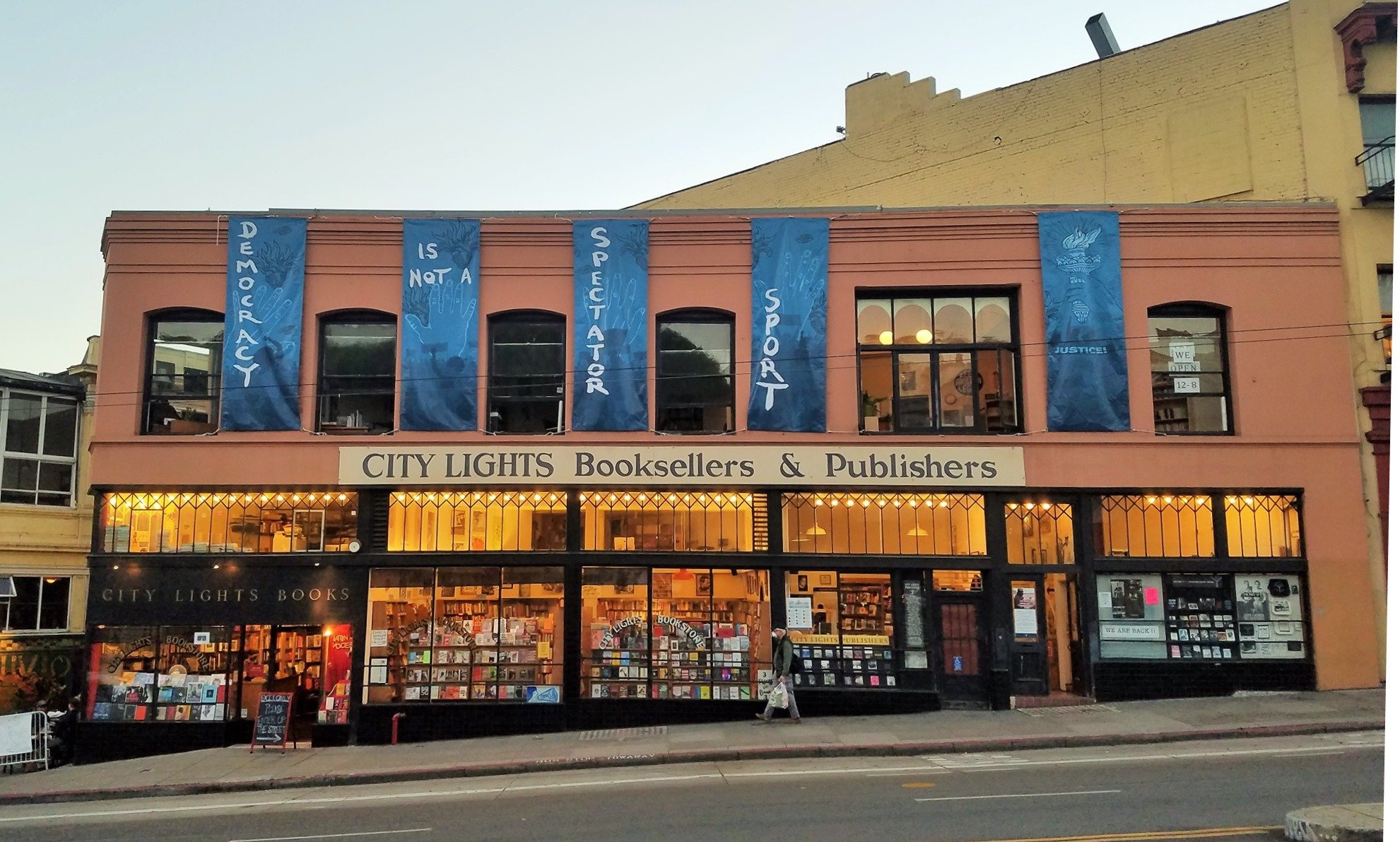 A square red building with two floors. There are five blue banners hanging from the top that read "Democracy is not a spectator sport." The inside is lit up with warm orange light.