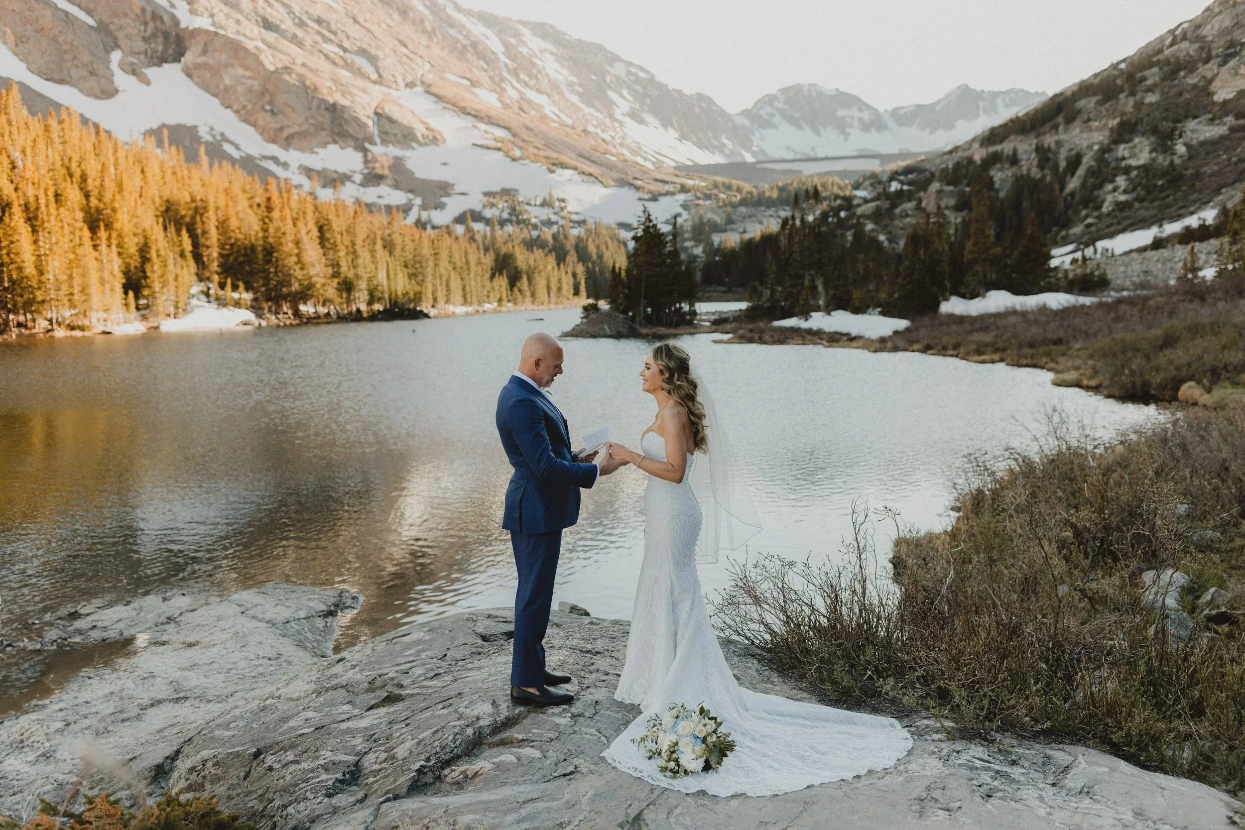 A wedding couple stands on a rocky shoreline by a lake in a mountainous area, with pine trees and snow-capped peaks in the background. The bride is in a white wedding dress with a flower bouquet on the ground, and the groom is in a blue suit. They are holding hands and exchanging vows.