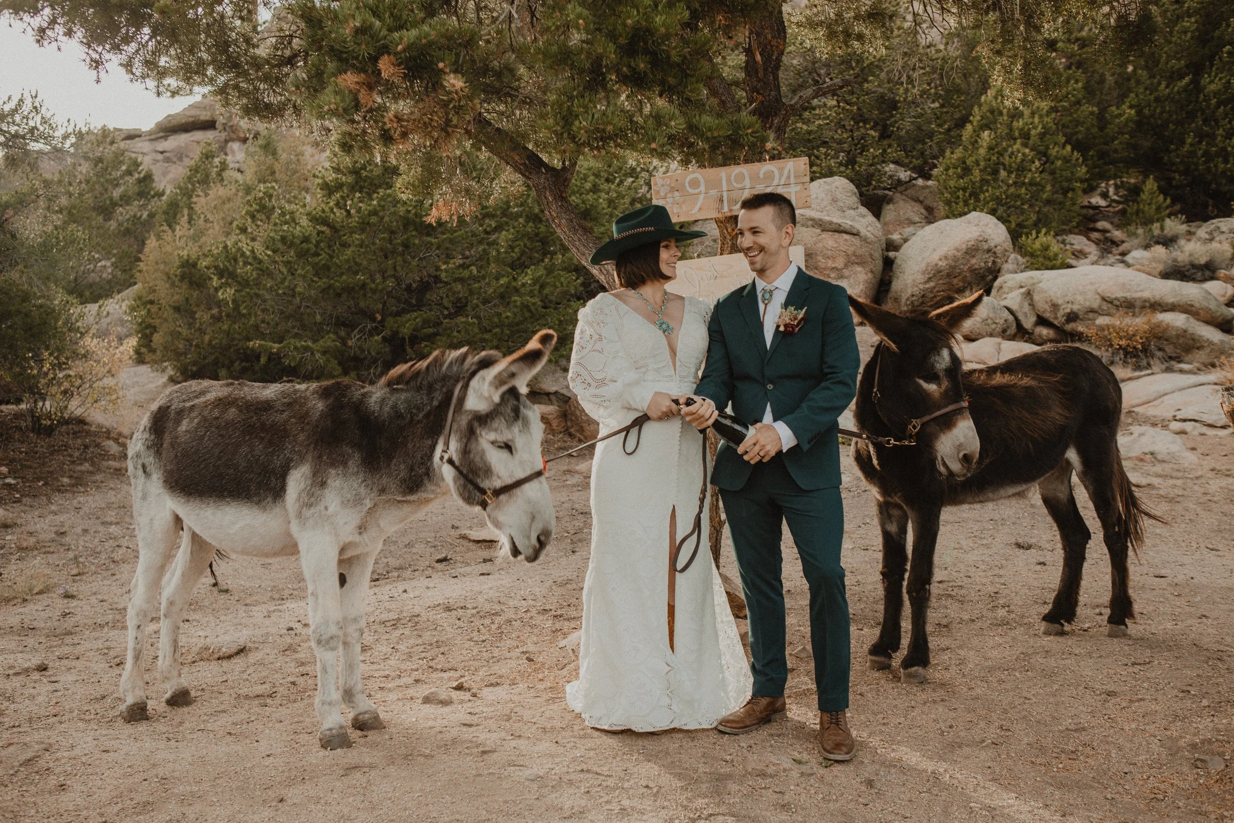 A man and woman dressed in wedding attire standing outdoors in a desert-like environment, holding a bottle of champagne, with two donkeys beside them and a tree behind. The woman wears a white dress with long sleeves and a large black hat, while the man wears a dark green suit. There is a wooden sign with the date 9-19-24 in the background.