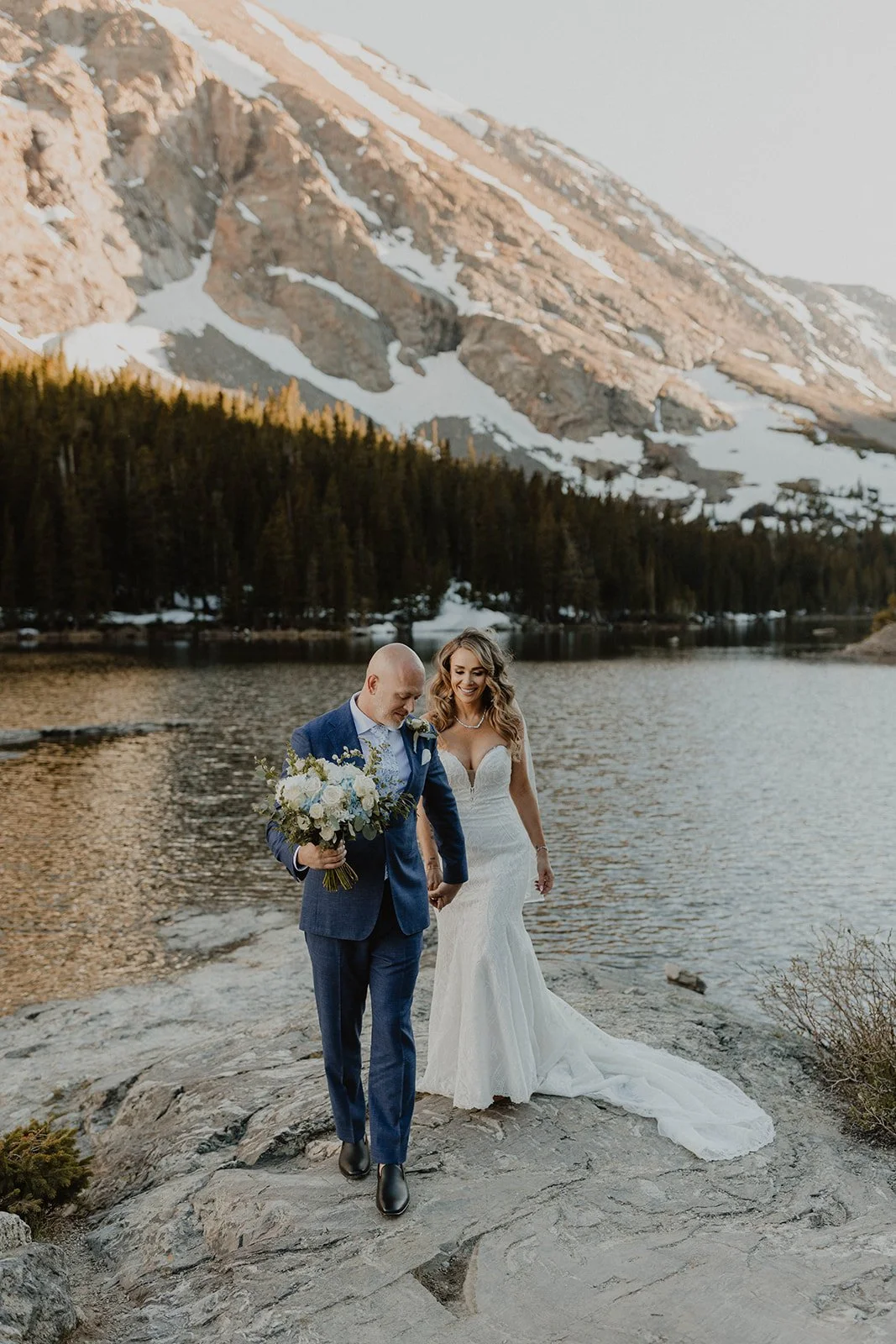 Bride and groom walking by a river in a mountainous landscape, with snow and trees in the background during sunset.