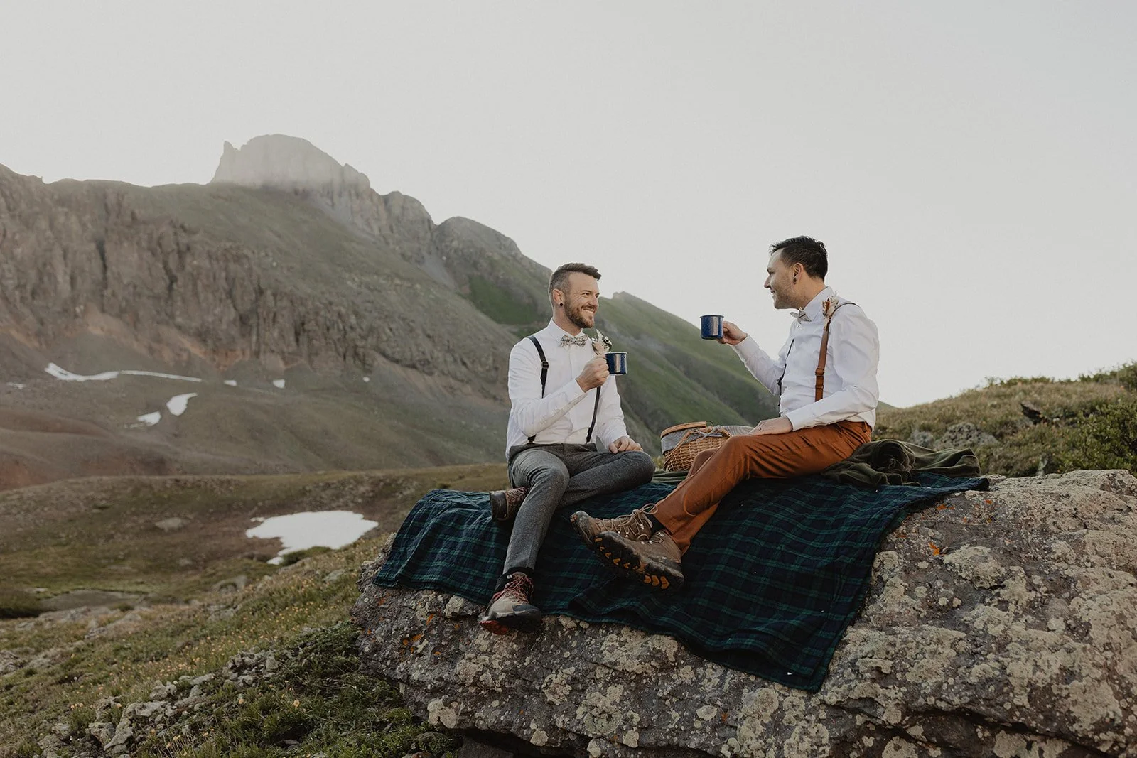 Two men sitting on a rock in a mountainous landscape, enjoying coffee, with patches of snow and green hills in the background.