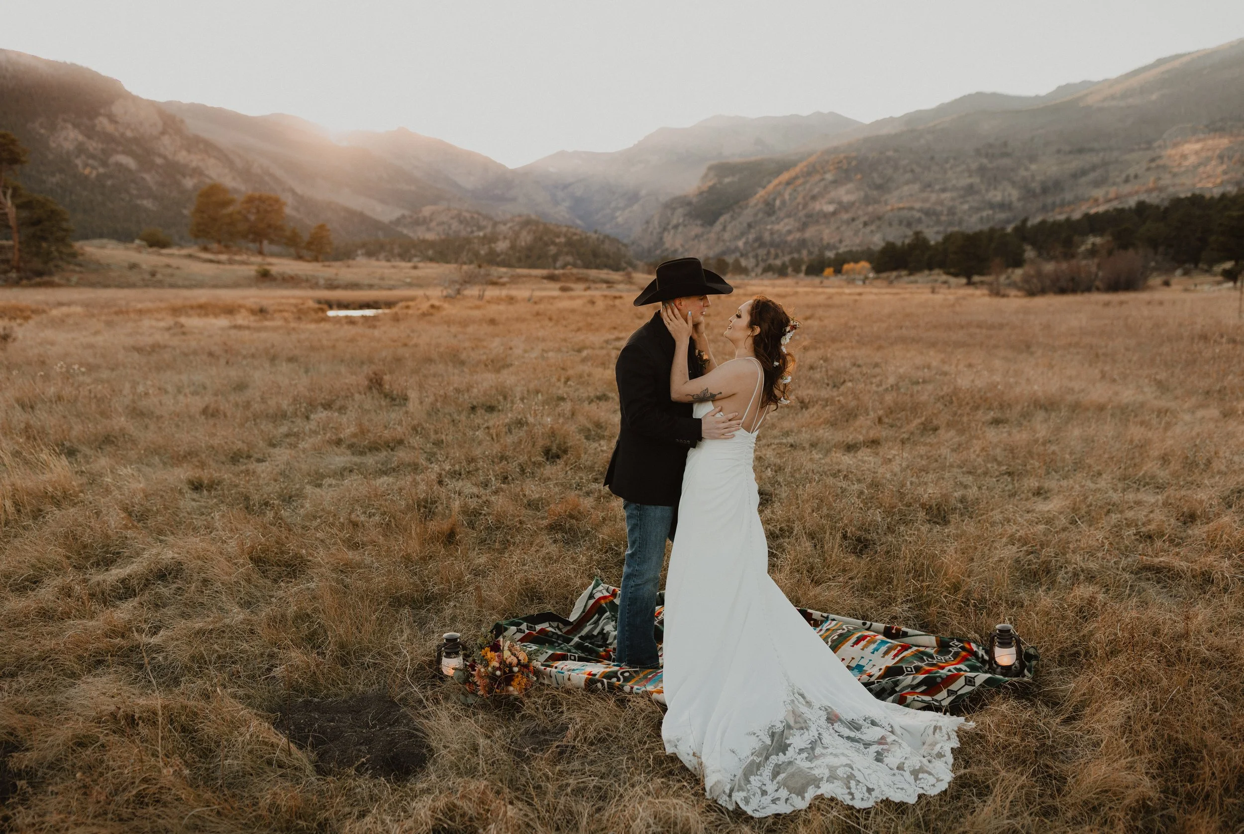 Couple in wedding attire standing on a patterned blanket in a field with mountains in the background, sunset lighting.