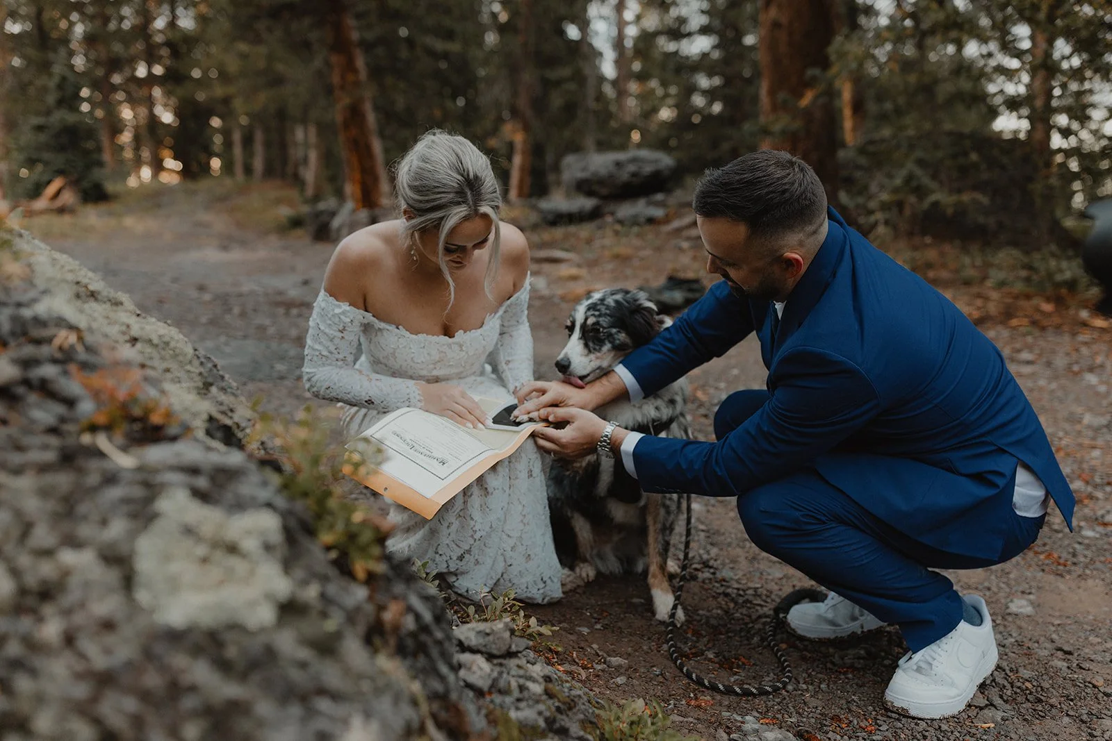A woman in a white wedding dress, a man in a blue suit, and a black and white dog, kneeling outdoors in a forest, with the woman signing a document.
