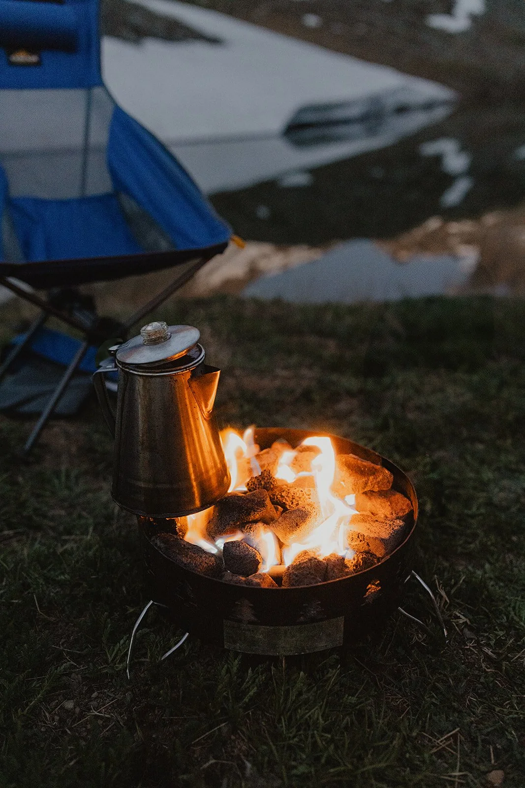 A camping scene with a small fire pit burning rocks and flames, with a metal kettle hanging over the fire, and camping chairs and a vehicle in the background.