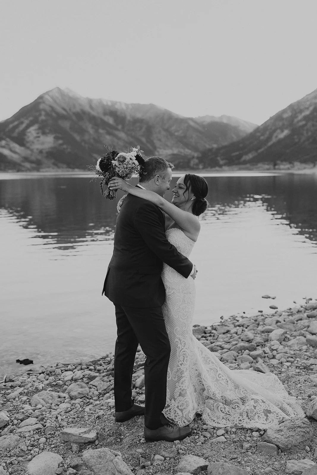 A bride and groom embrace on a rocky lakeshore, with mountains and a lake in the background, in a black and white wedding photo.