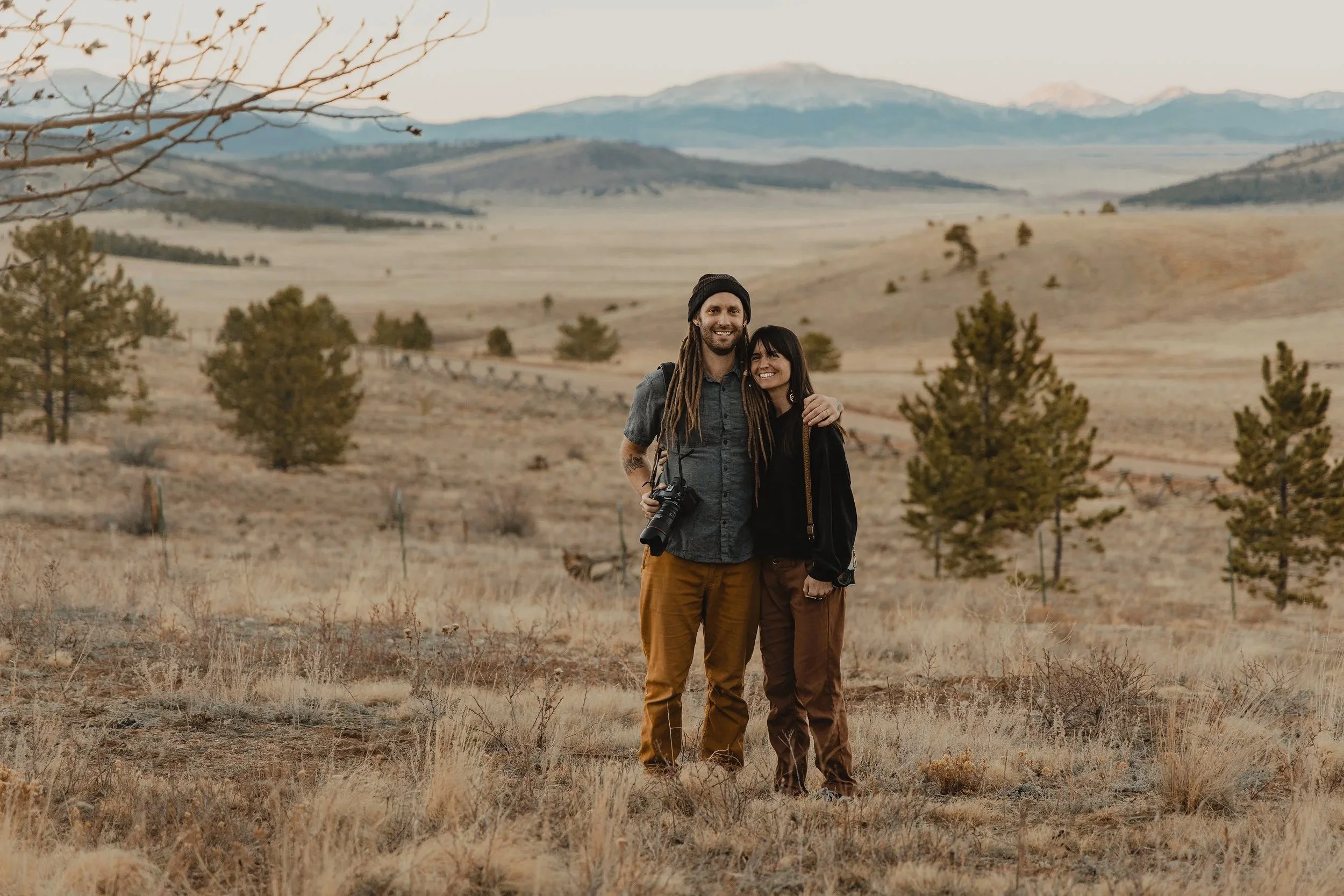 a married couple elopement photographers standing in a meadow with mountain views in the background at sunset