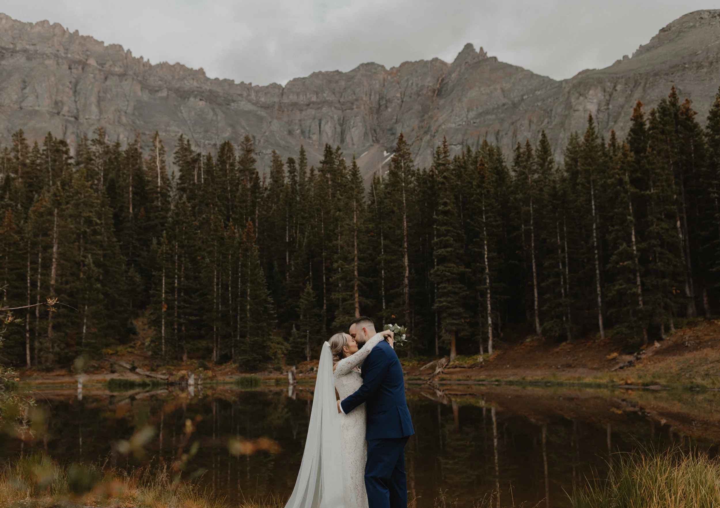 A bride and groom sharing a kiss by a lake with mountains and pine trees in the background on a cloudy day.