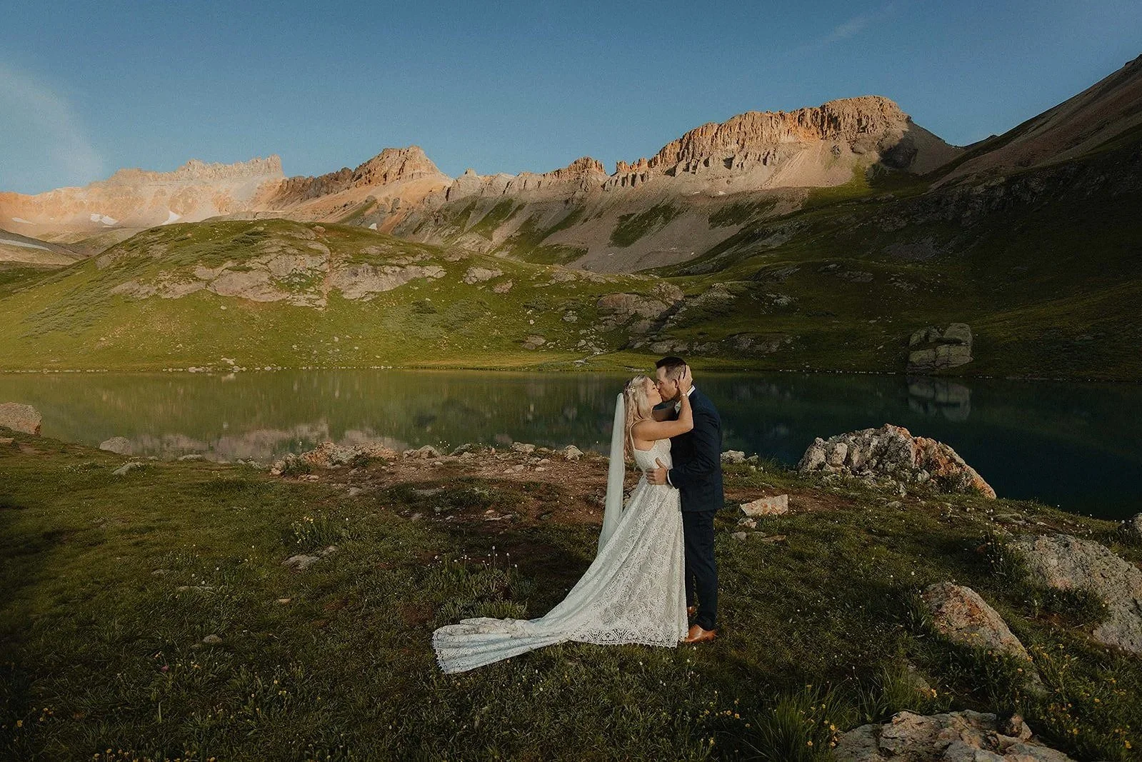 A bride and groom share a kiss by a mountain lake in a scenic outdoor setting with mountains and green hills in the background.
