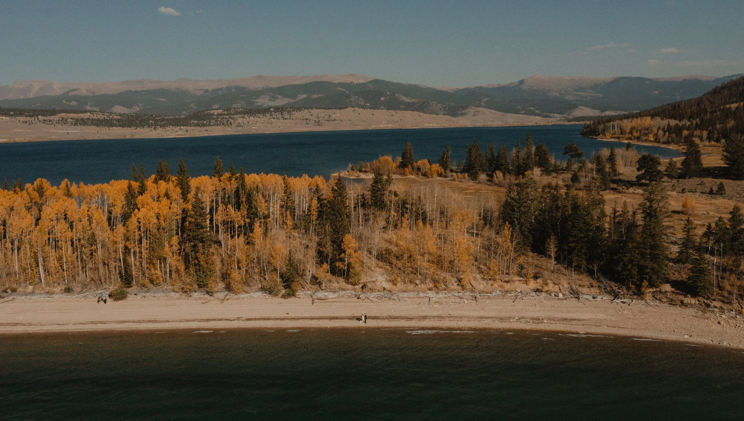 Aerial view of a lakeside with a sandy beach, a wooded area with fall foliage, and mountains in the background under a clear blue sky.