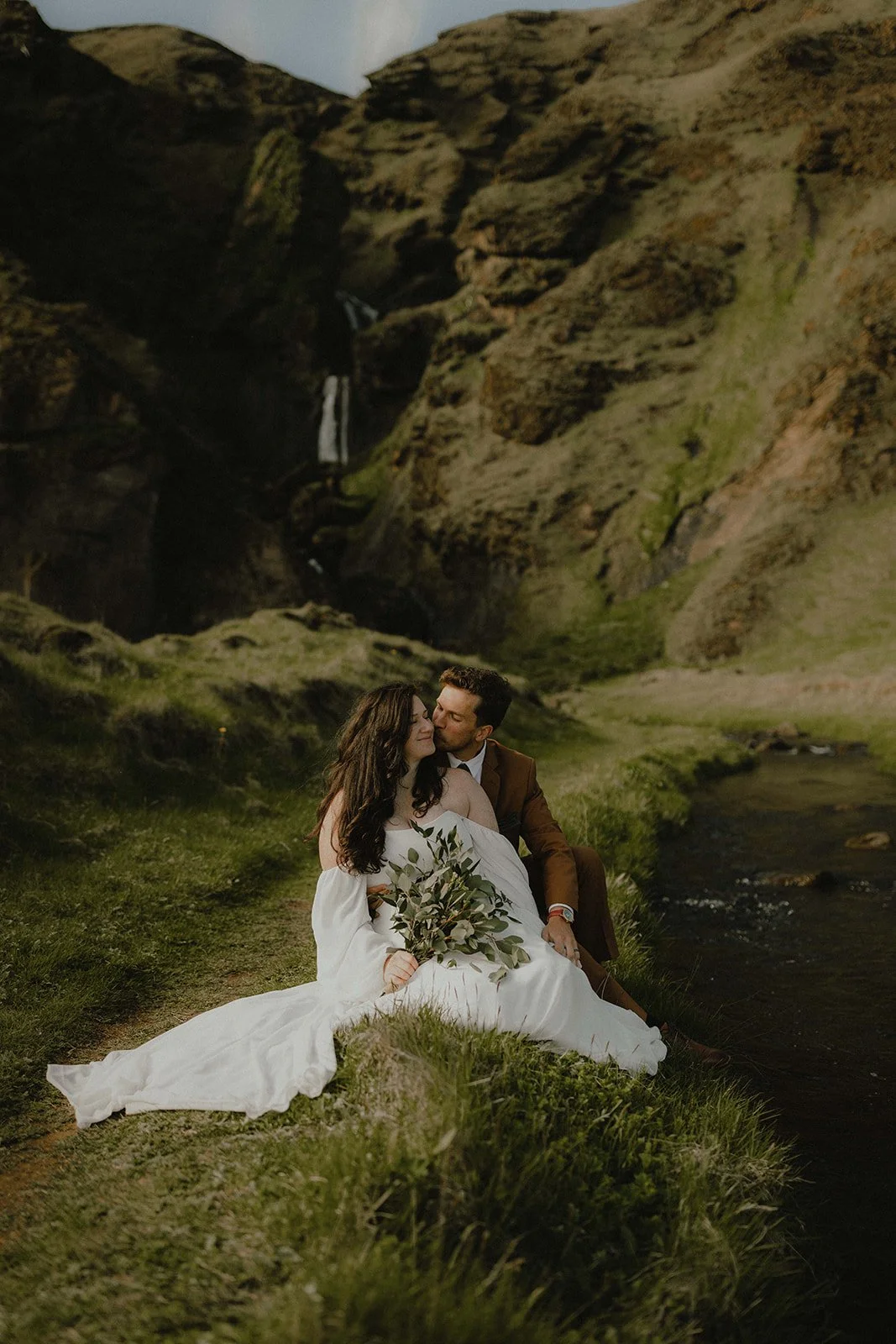 A couple sits on the grass near a stream in a lush green landscape with steep cliffs and a waterfall in the background, during a romantic moment.