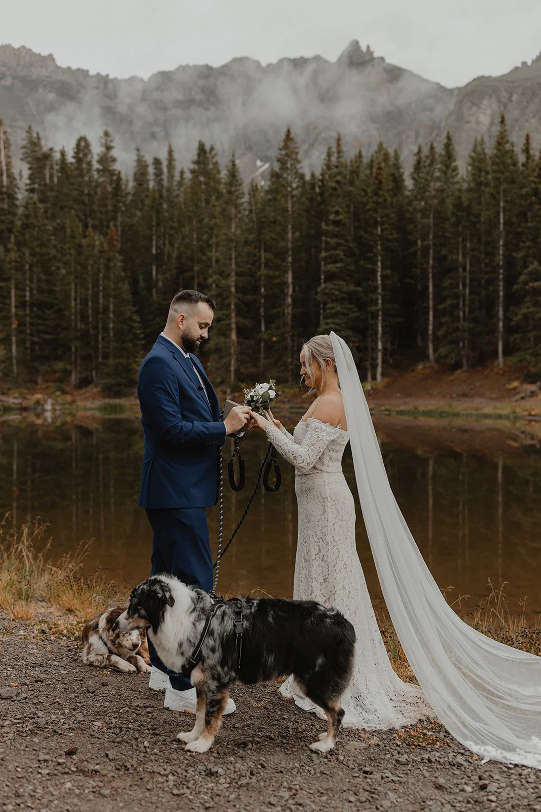 A bride and groom exchanging vows outdoors near a lake with a mountain and forest background, accompanied by a dog.