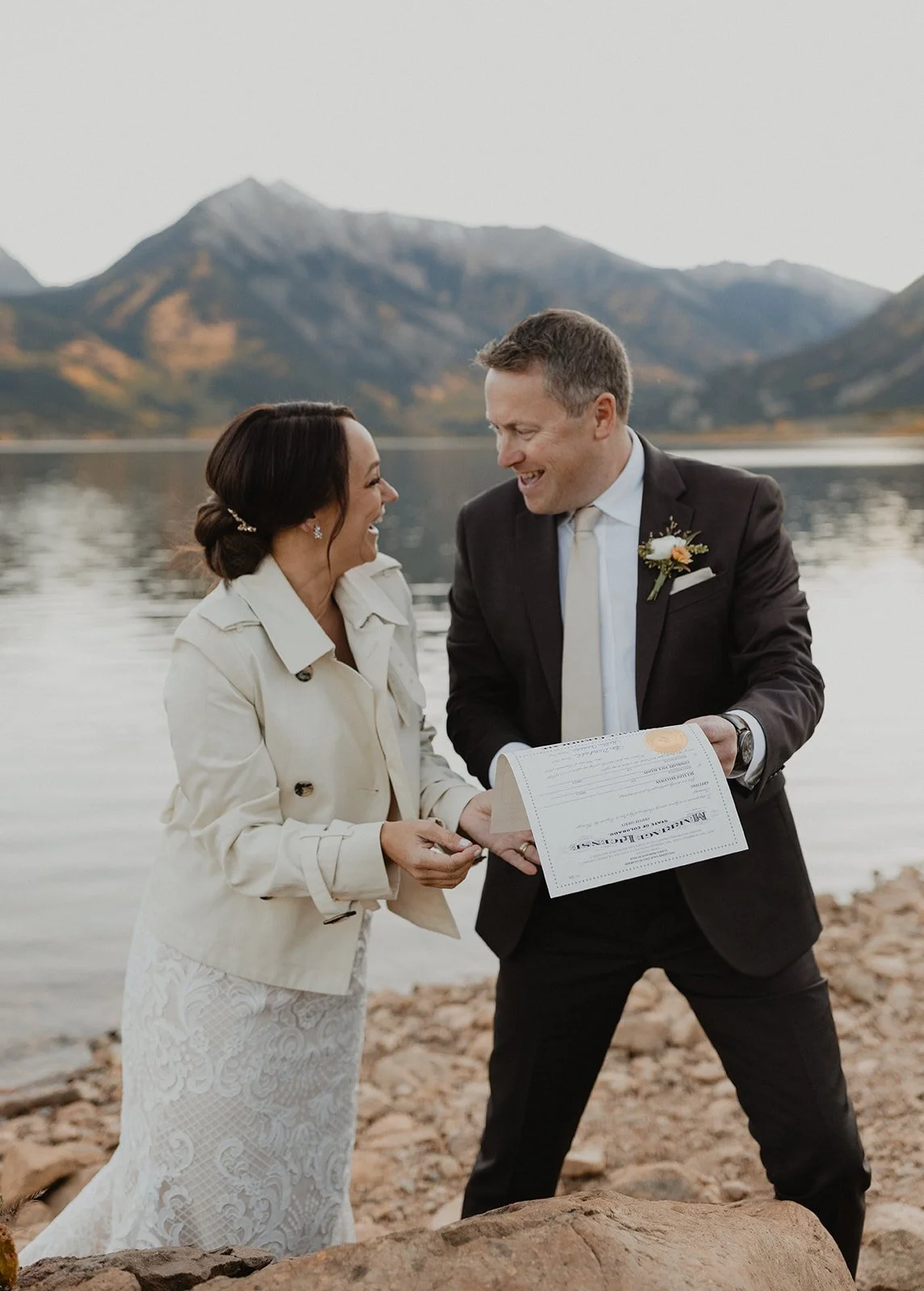 A couple dressed in wedding attire exchanging vows by a lake with mountains in the background.