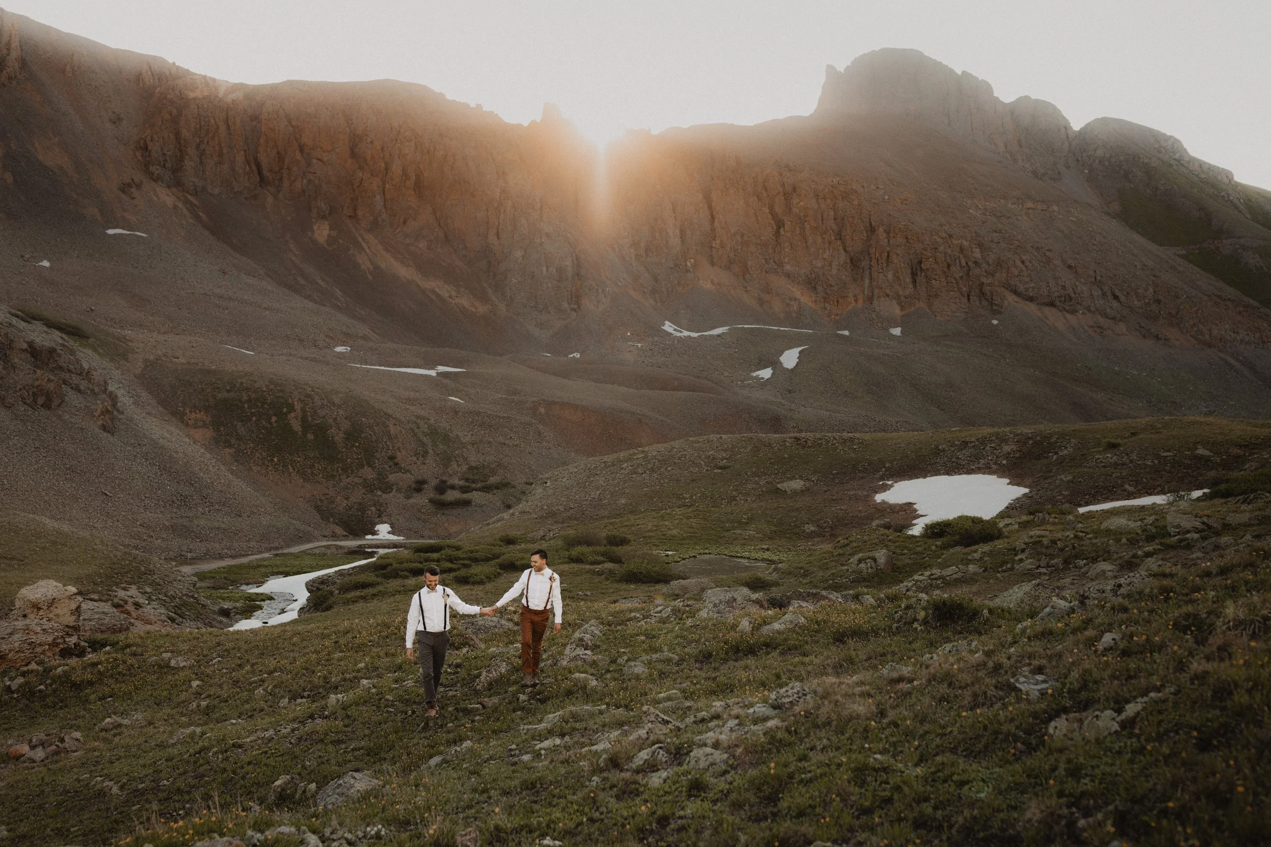 Two men on their elopement day in Silverton, Colorado walking hand-in-hand across a rugged, grassy landscape with small patches of snow, surrounded by mountains at sunset.