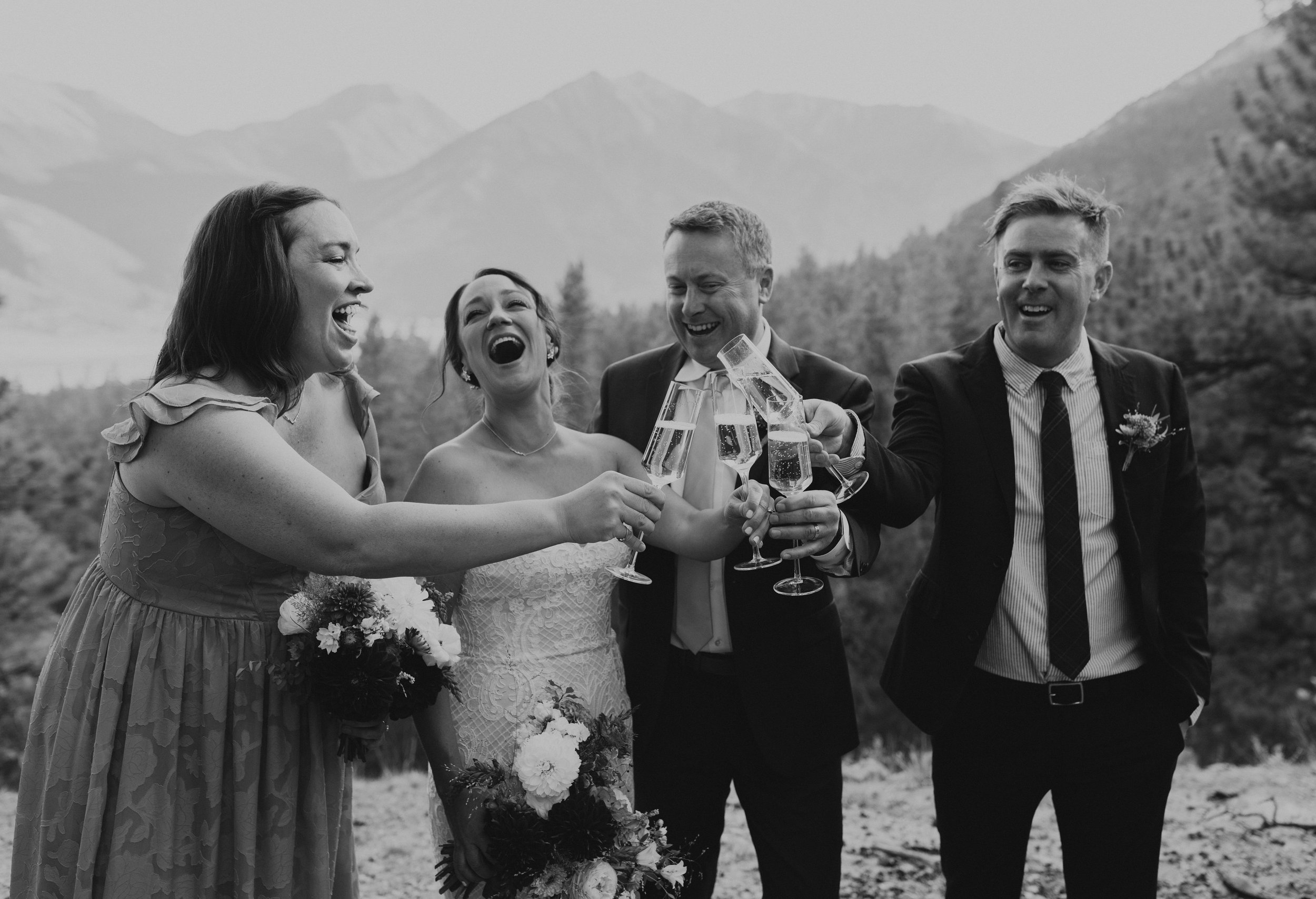A group of five people, including two women in wedding dresses and three men in suits, celebrating with cheers and glasses of champagne outdoors with mountains in the background.