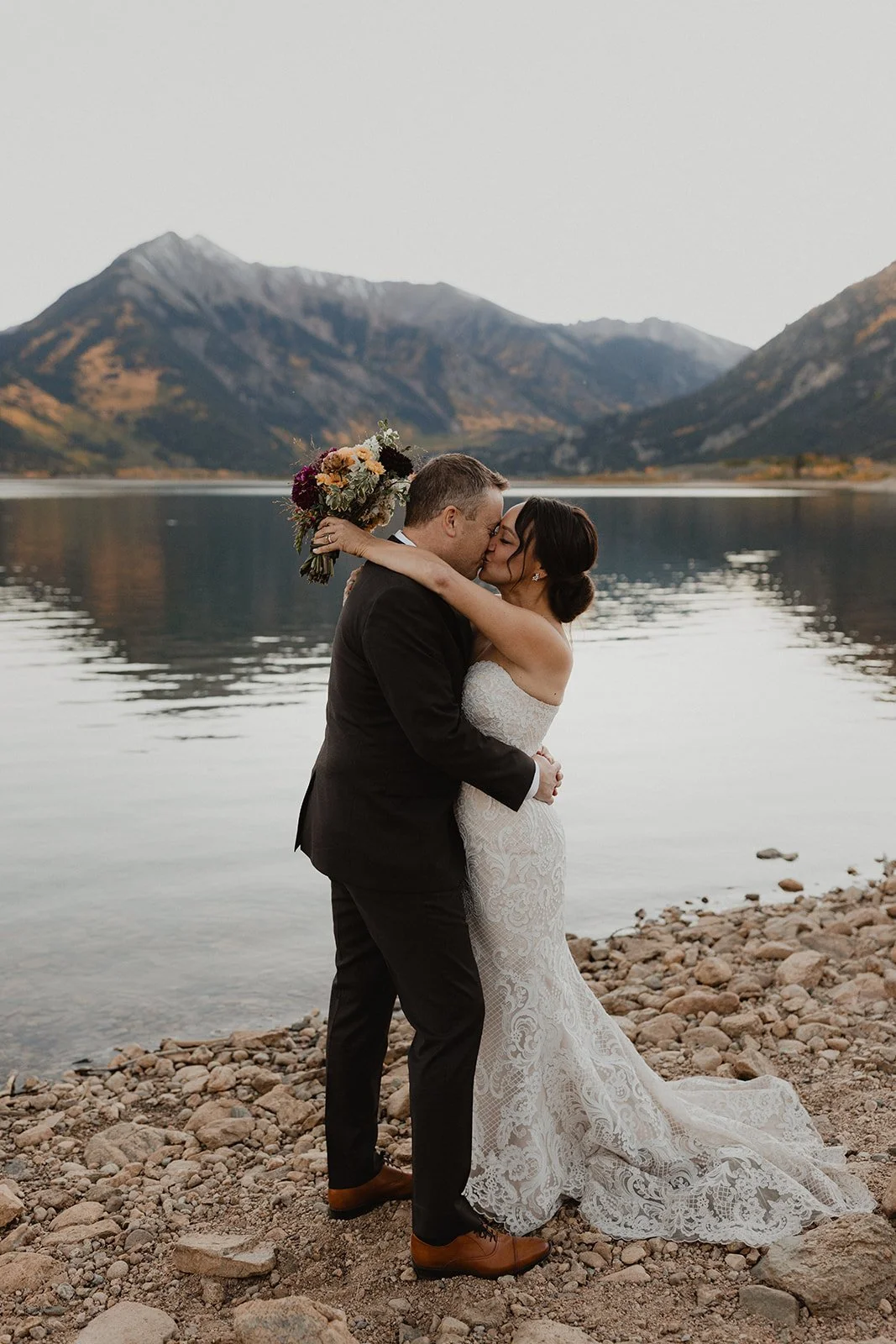 A newlywed couple embracing and kissing by a lake with mountains in the background, the bride holding a bouquet of flowers.