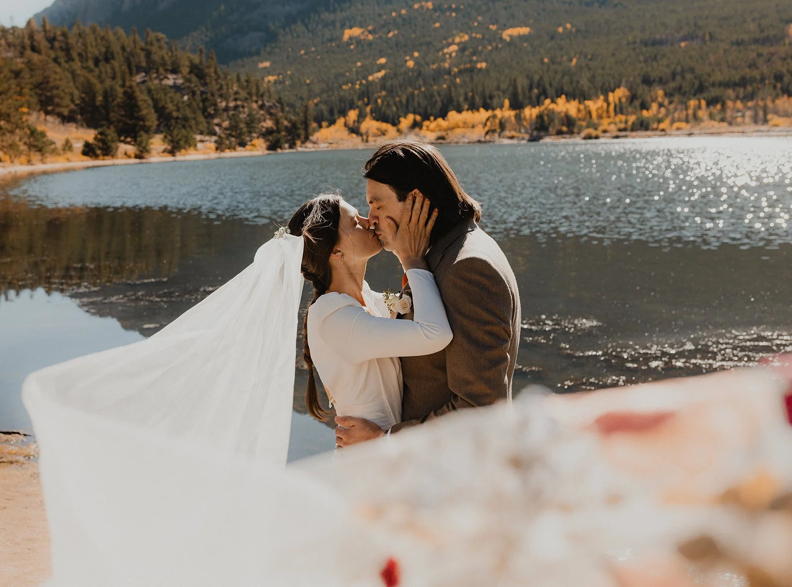 A bride and groom sharing a kiss by a lake with mountains and autumn trees in the background.