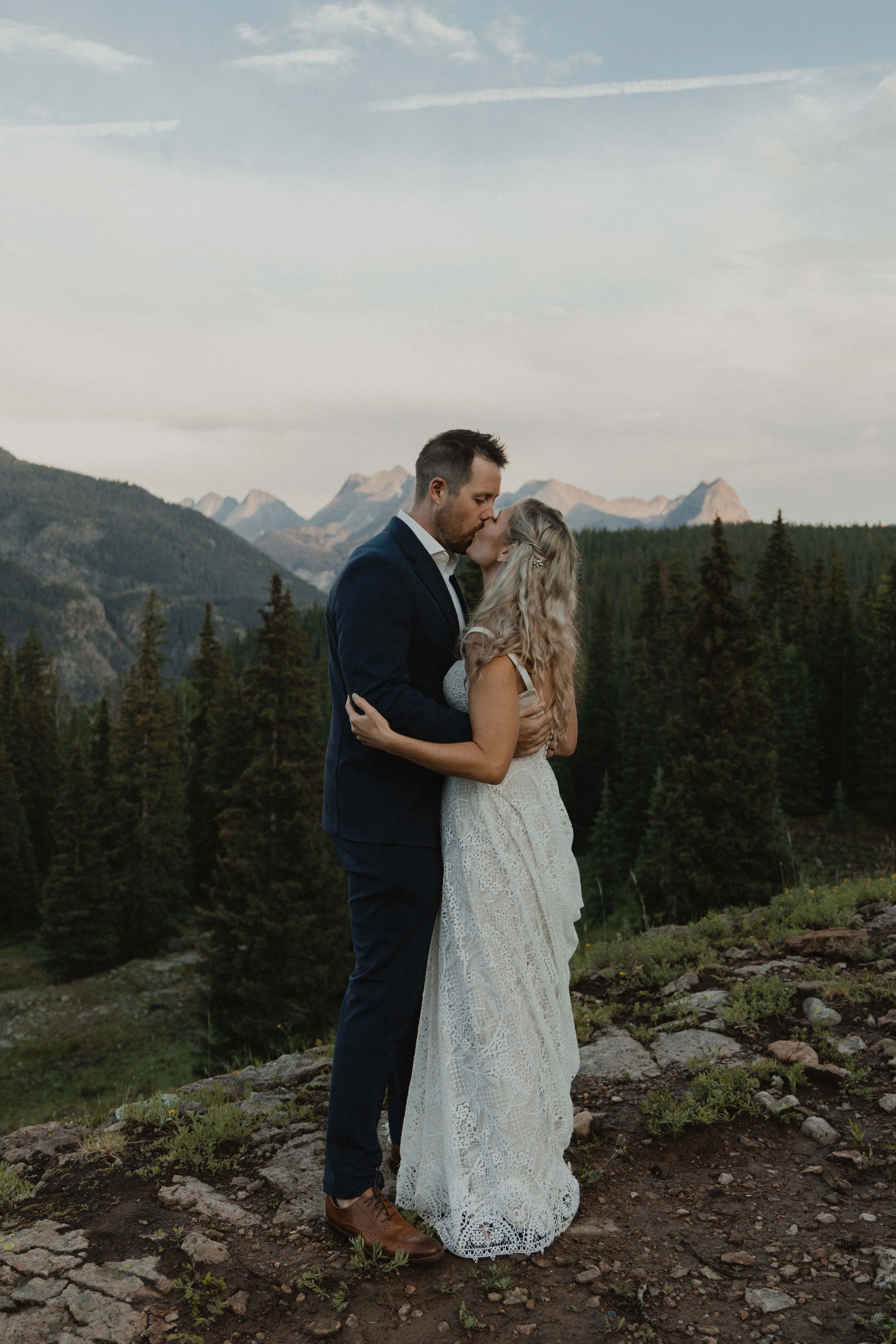 A couple dressed in wedding attire sharing a kiss outdoors on rocky terrain with a mountain and forest background.
