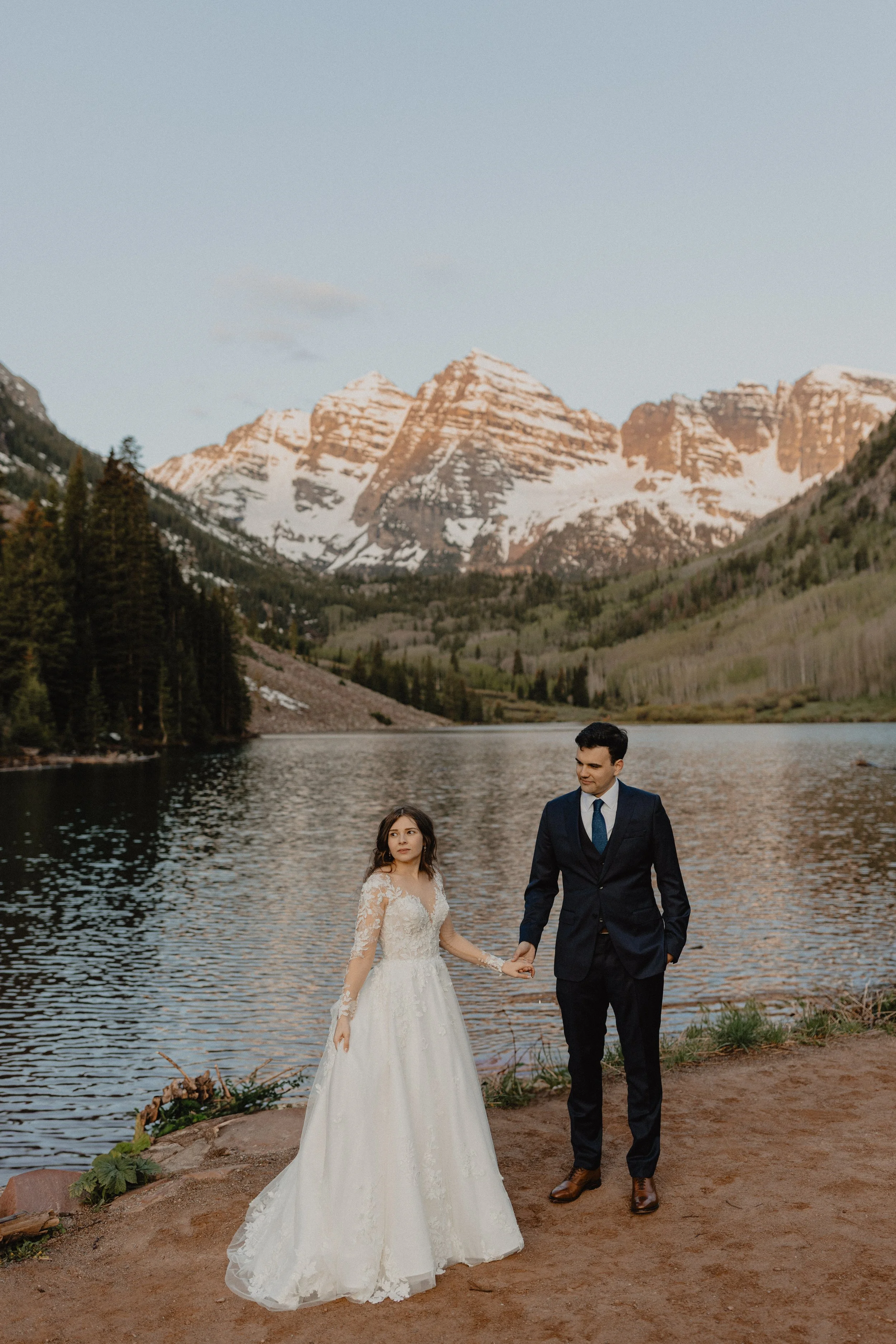A bride in a white wedding dress and a groom in a dark suit holding hands near a lake with Maroon Bells in the background.