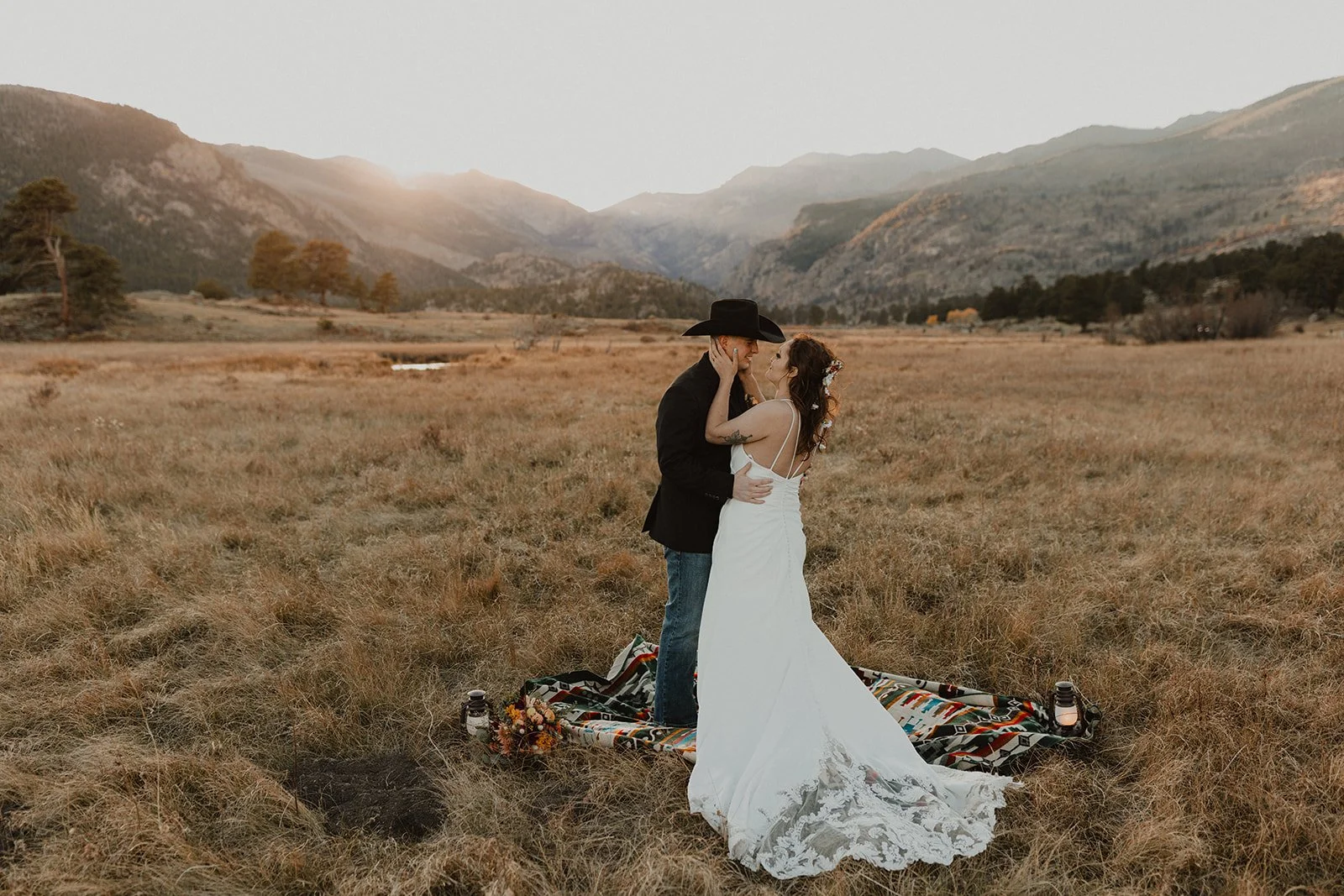 A couple in wedding attire stands close together on a patterned blanket in a field during sunset, with mountains and a partly cloudy sky in the background.