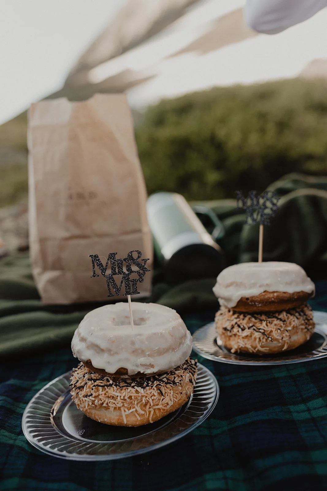 Two donuts topped with white glaze and sprinkles, each on a small plate, with 'Mrs & Mr' cake toppers, and a paper bag in the background on a green plaid cloth.