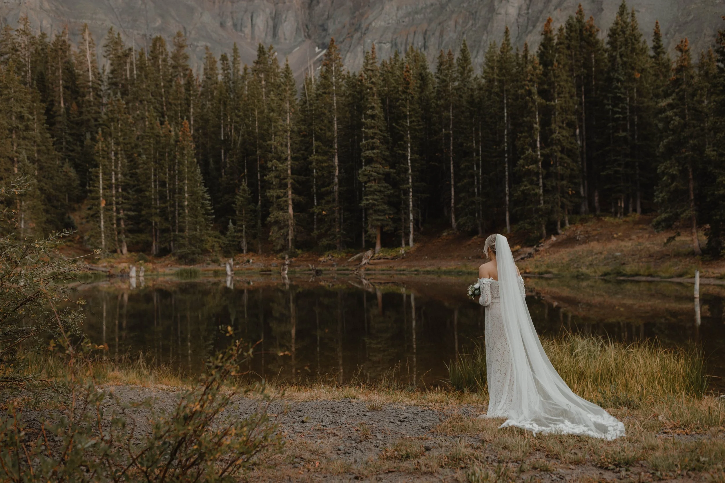 A bride in a white lace wedding gown with a long train and veil stands by a lake at the edge of a forest in Telluride, Colorado. She is holding a bouquet, with mountains in the background.