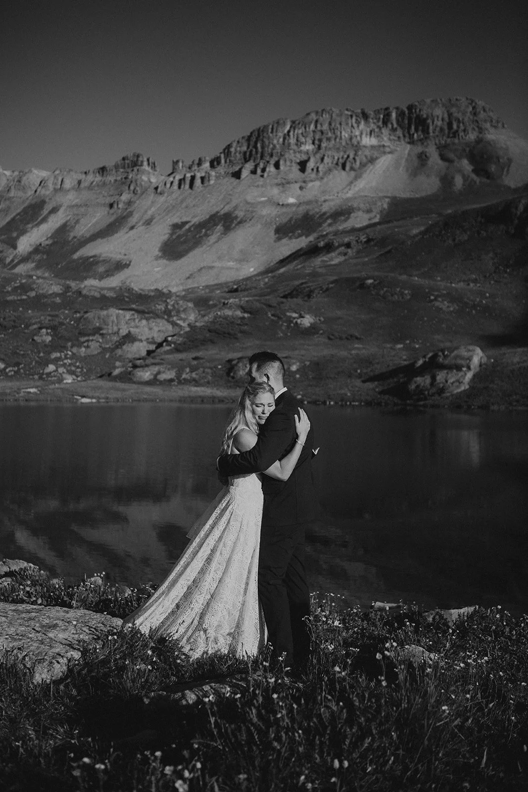 A black-and-white photo of a couple hugging outdoors near a lake in Silverton, Colorado with mountains in the background.