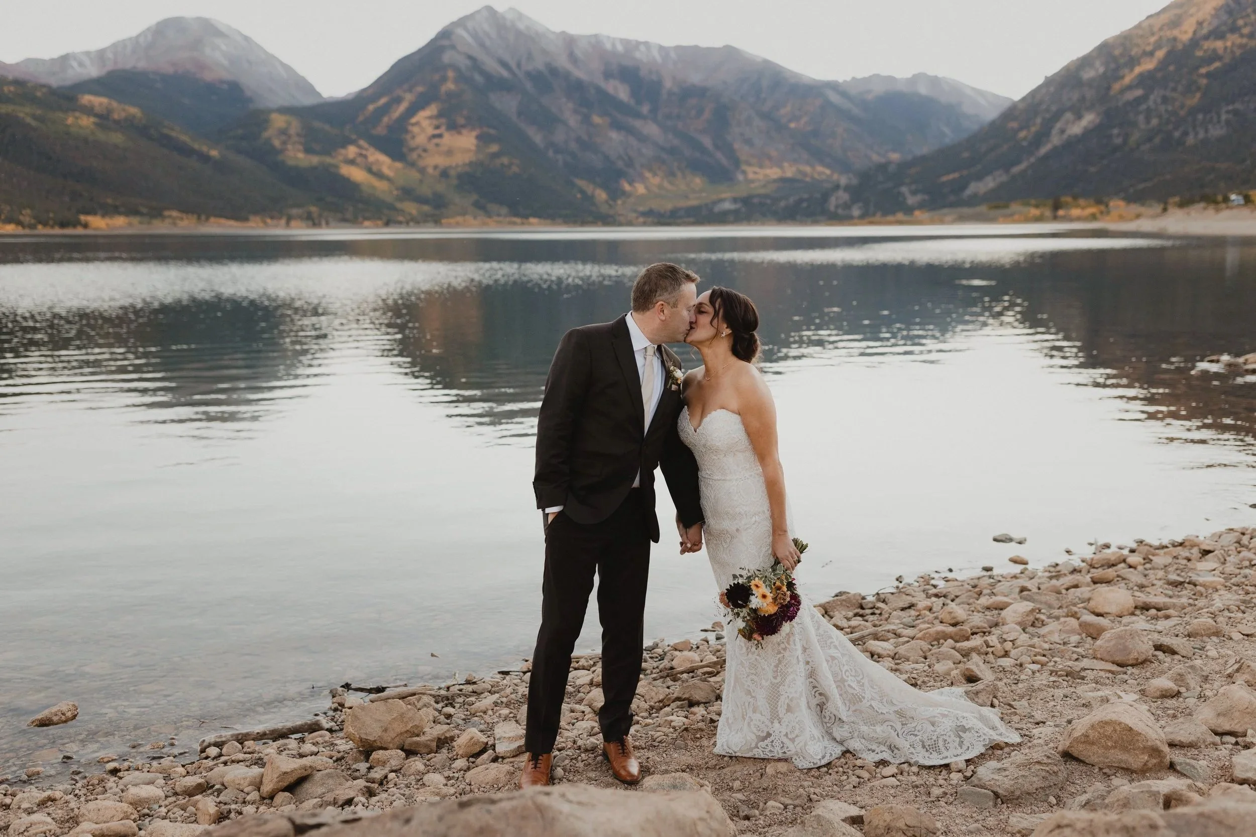 A newlywed couple sharing a kiss by a lake, with mountains in the background, during sunset.