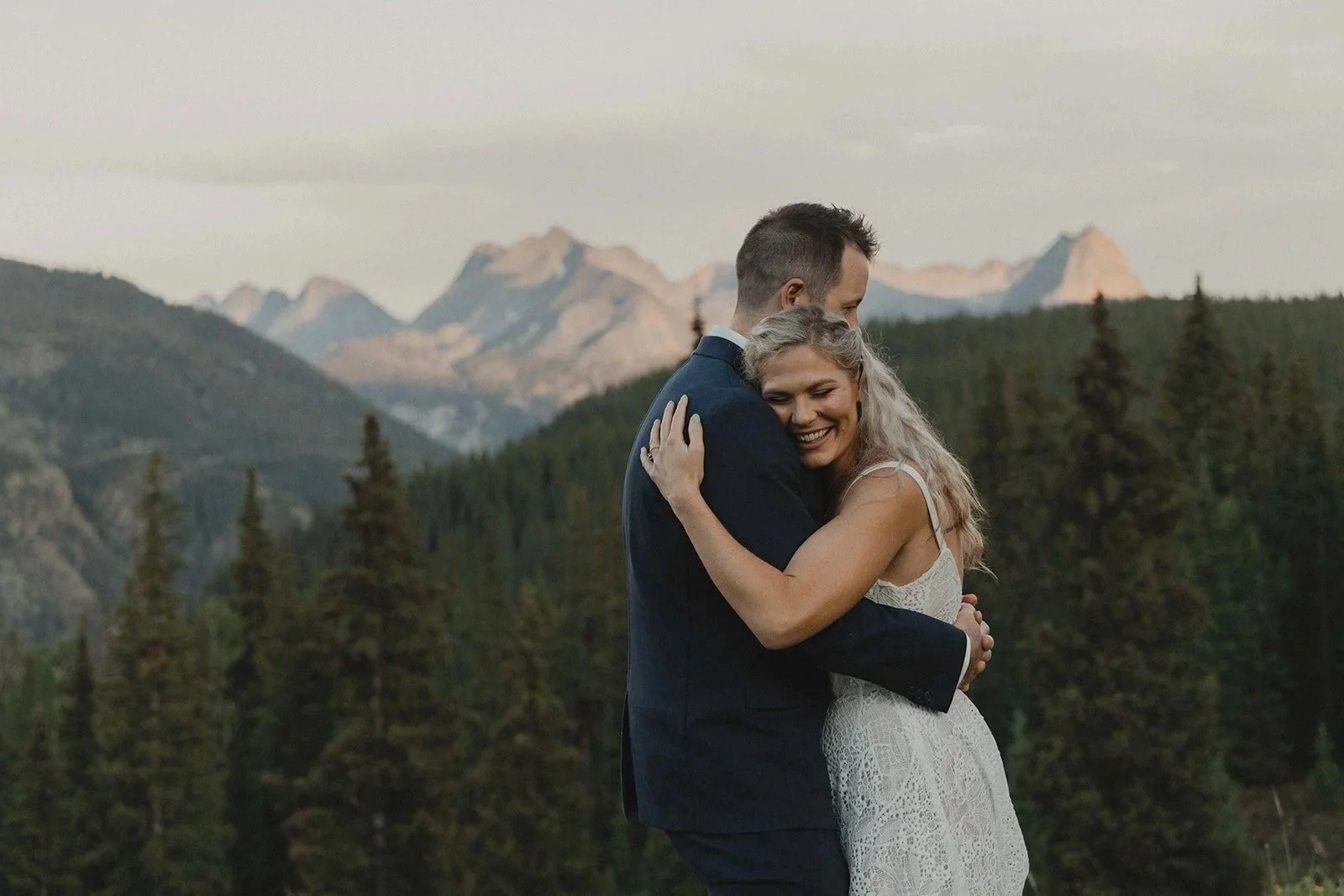 A couple embraces outdoors in front of mountain scenery, with the woman smiling and the man holding her close.