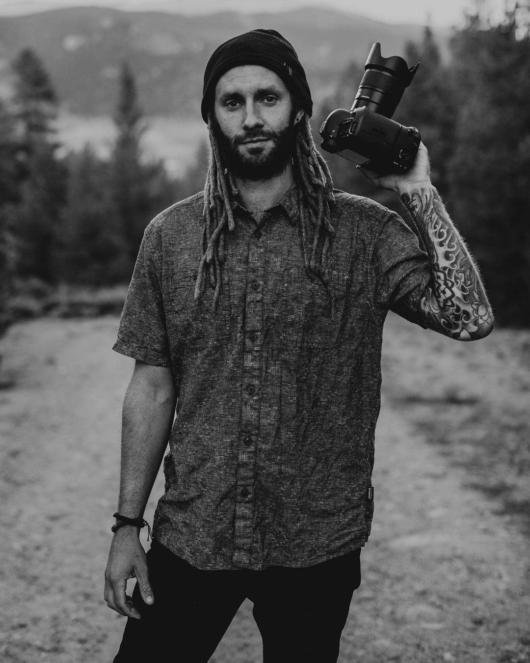 A man with dreadlocks, wearing a beanie, holding a professional camera on his shoulder, standing outdoors in a natural landscape.