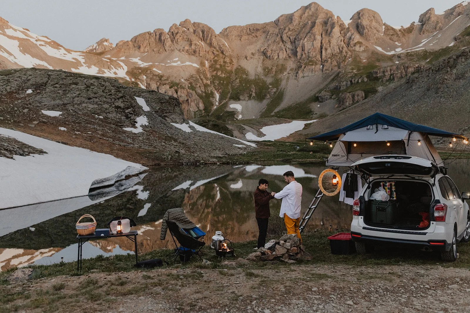 Two men stand near a campsite by a lake in a mountainous area during sunset. The campsite has a car with a rooftop tent decorated with string lights and a small table with lanterns and supplies. The background features snow-capped mountains reflected in the lake.