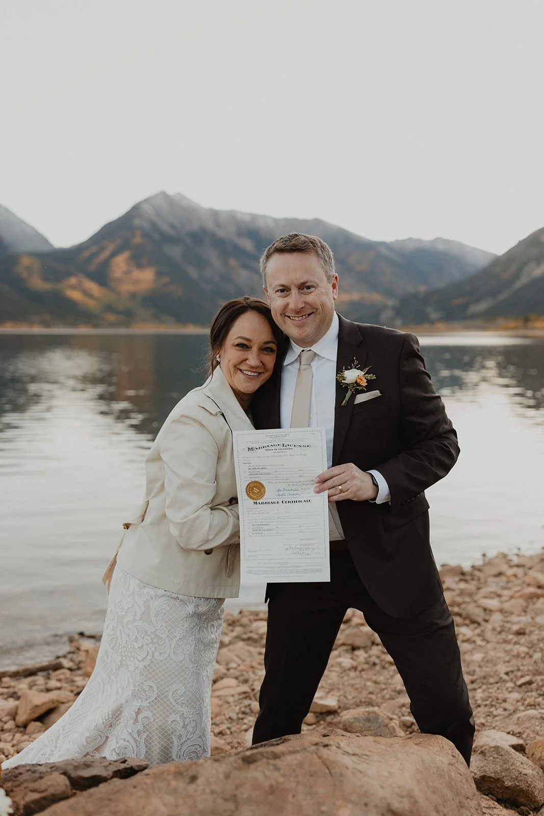 A smiling couple holding a marriage certificate, standing on a rocky shoreline with a lake and mountains in the background.