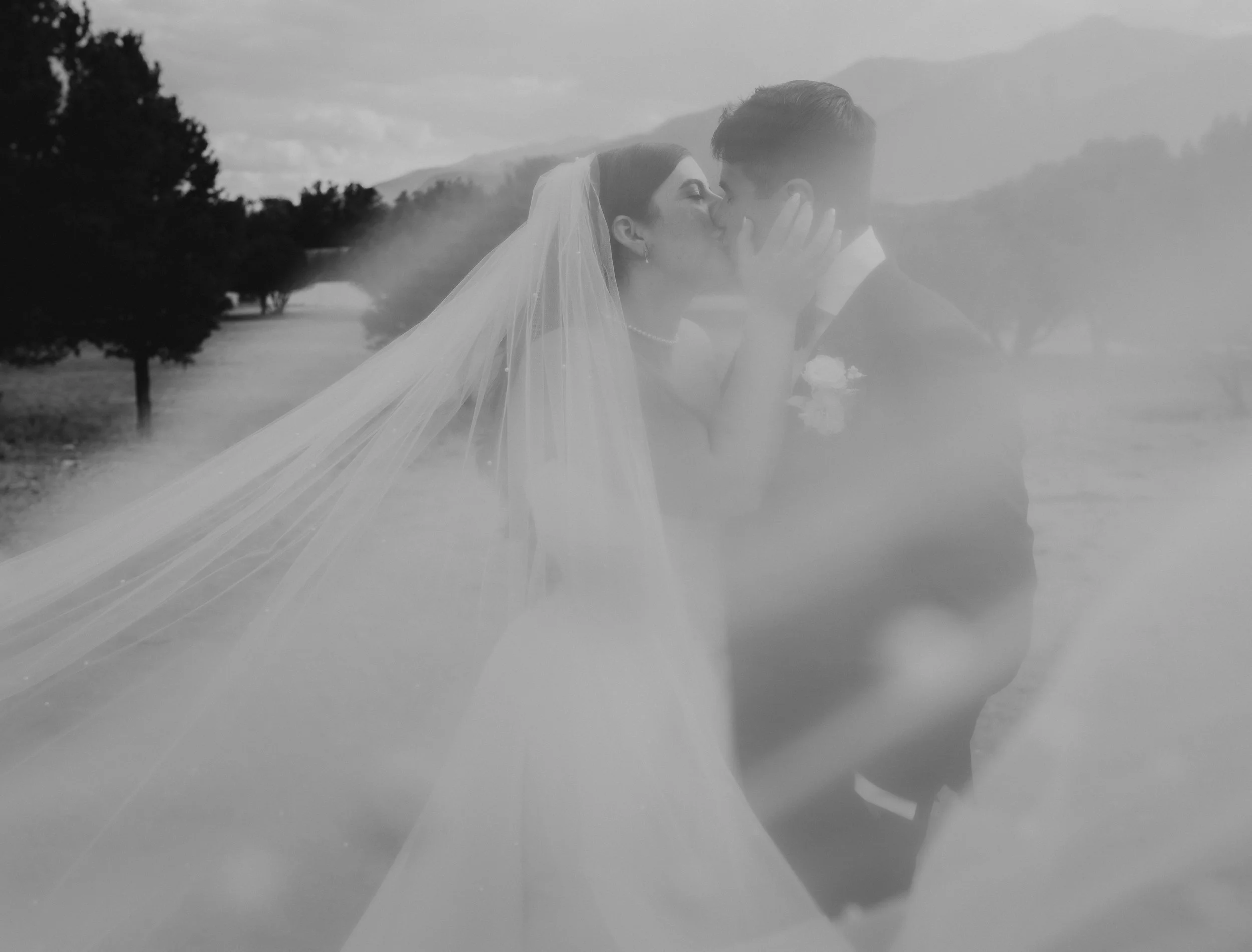 Black and white photo of a bride and groom sharing a kiss outdoors, with trees and mountains in the background, with a foggy or smoky effect partially obscuring the image.