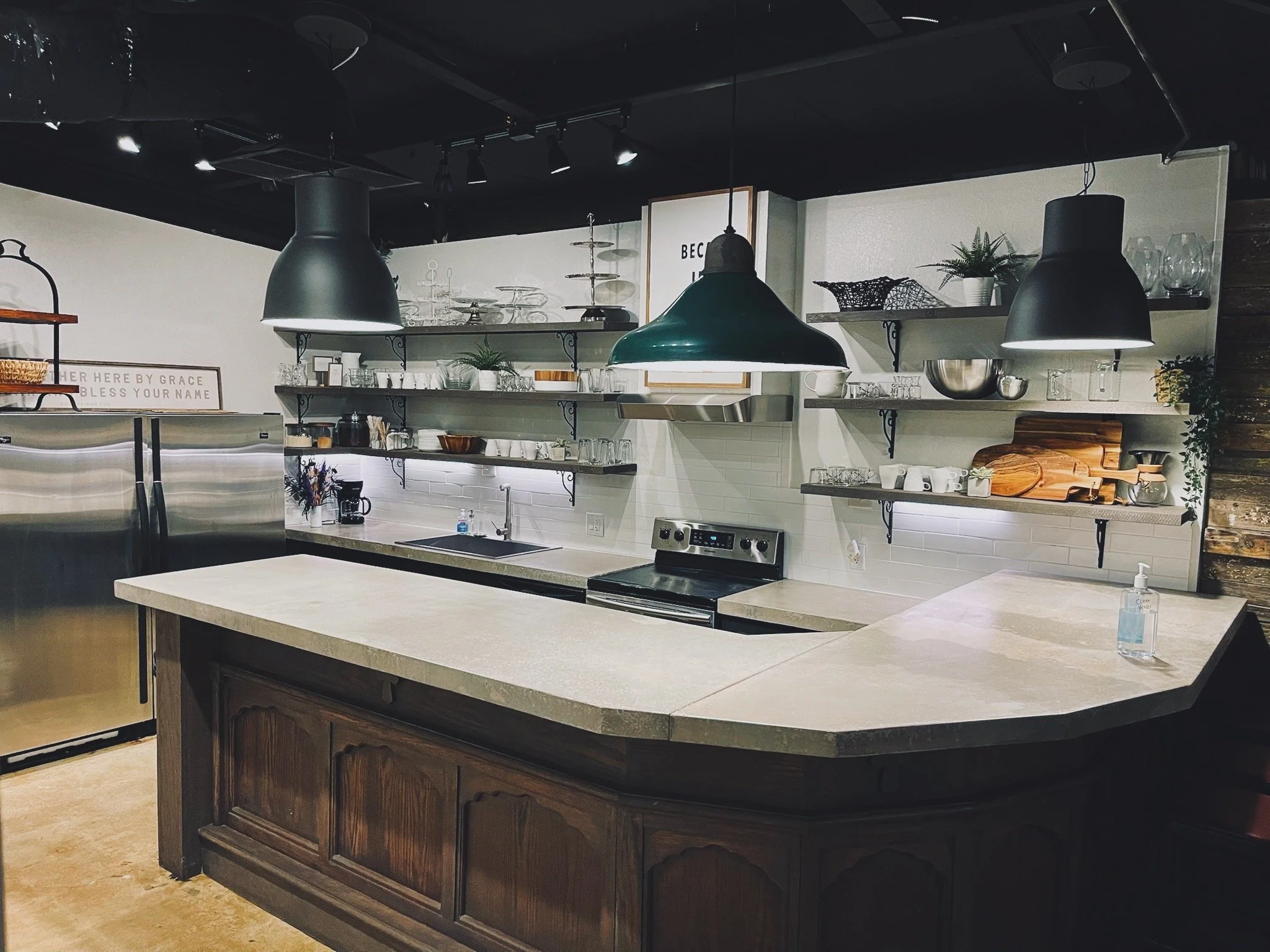Modern kitchen with open shelving, black pendant lights, stainless steel refrigerator, and a central island with a dark wood base and concrete countertop.