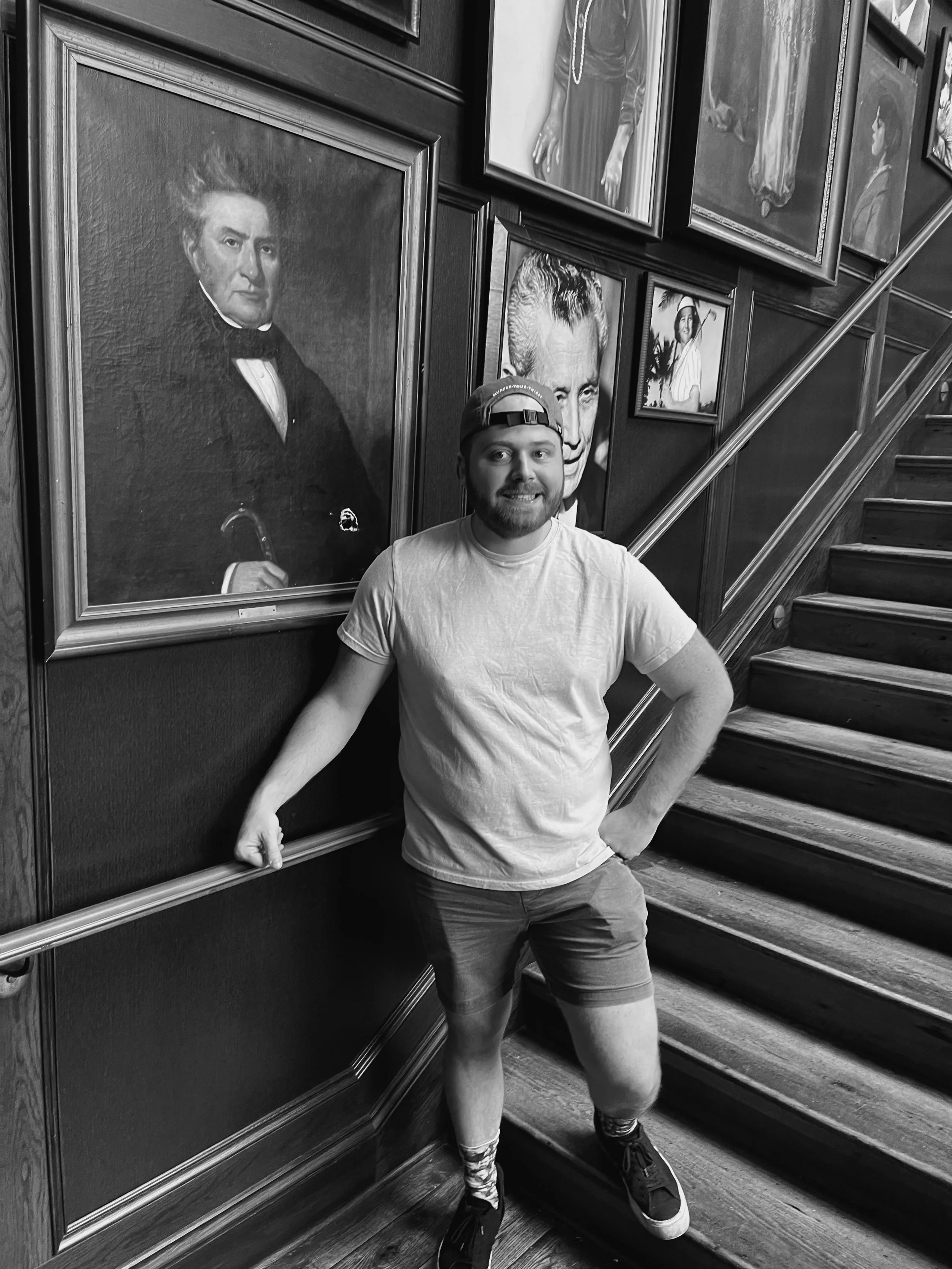 A man wearing a cap, t-shirt, shorts, and sneakers standing on a staircase, smiling at the camera. Portraits of historical figures are hanging on the wall behind him.