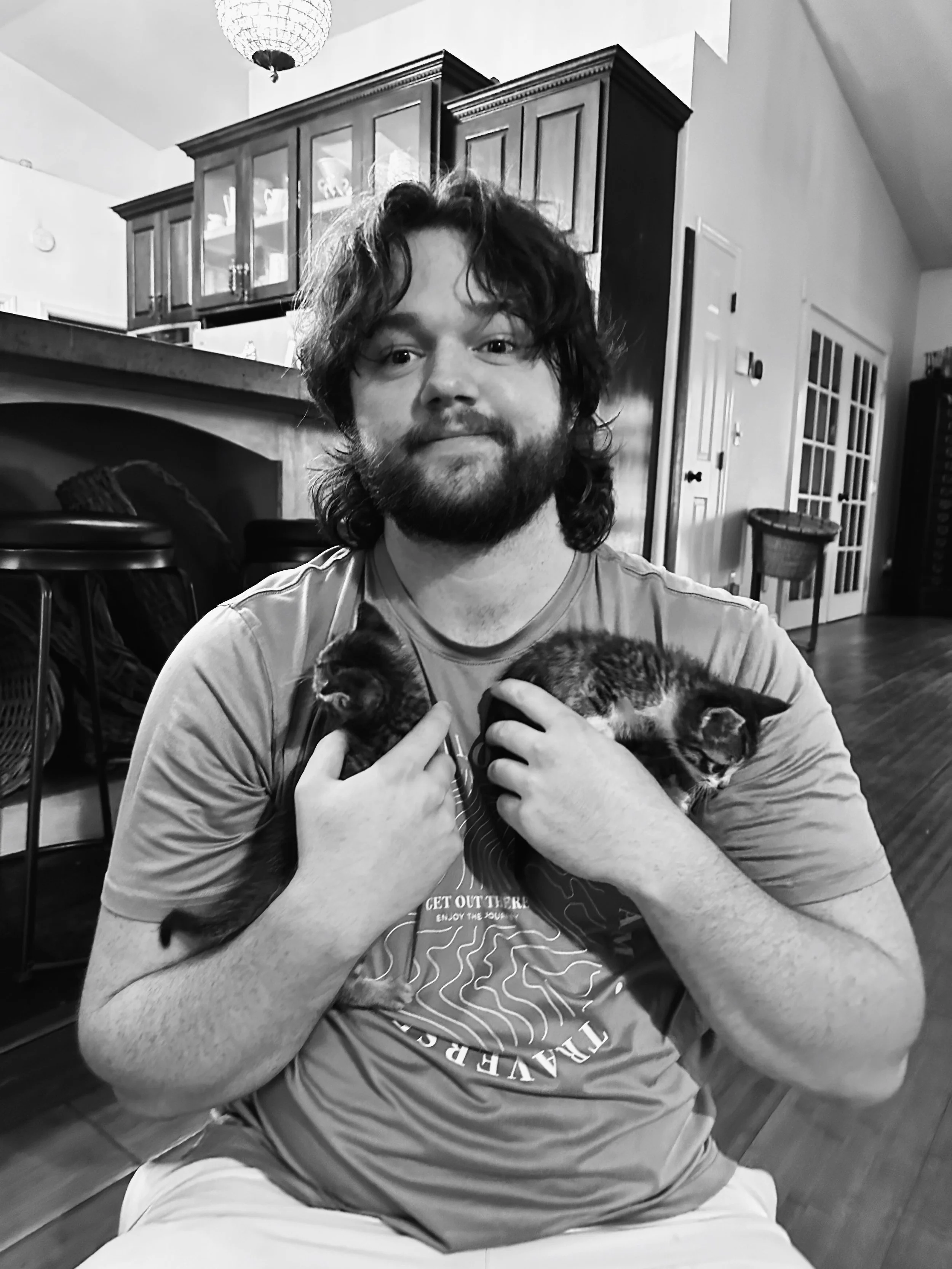 A man with curly hair and a beard holding two kittens, one in each arm, inside a home with wooden furniture and a kitchen in the background.