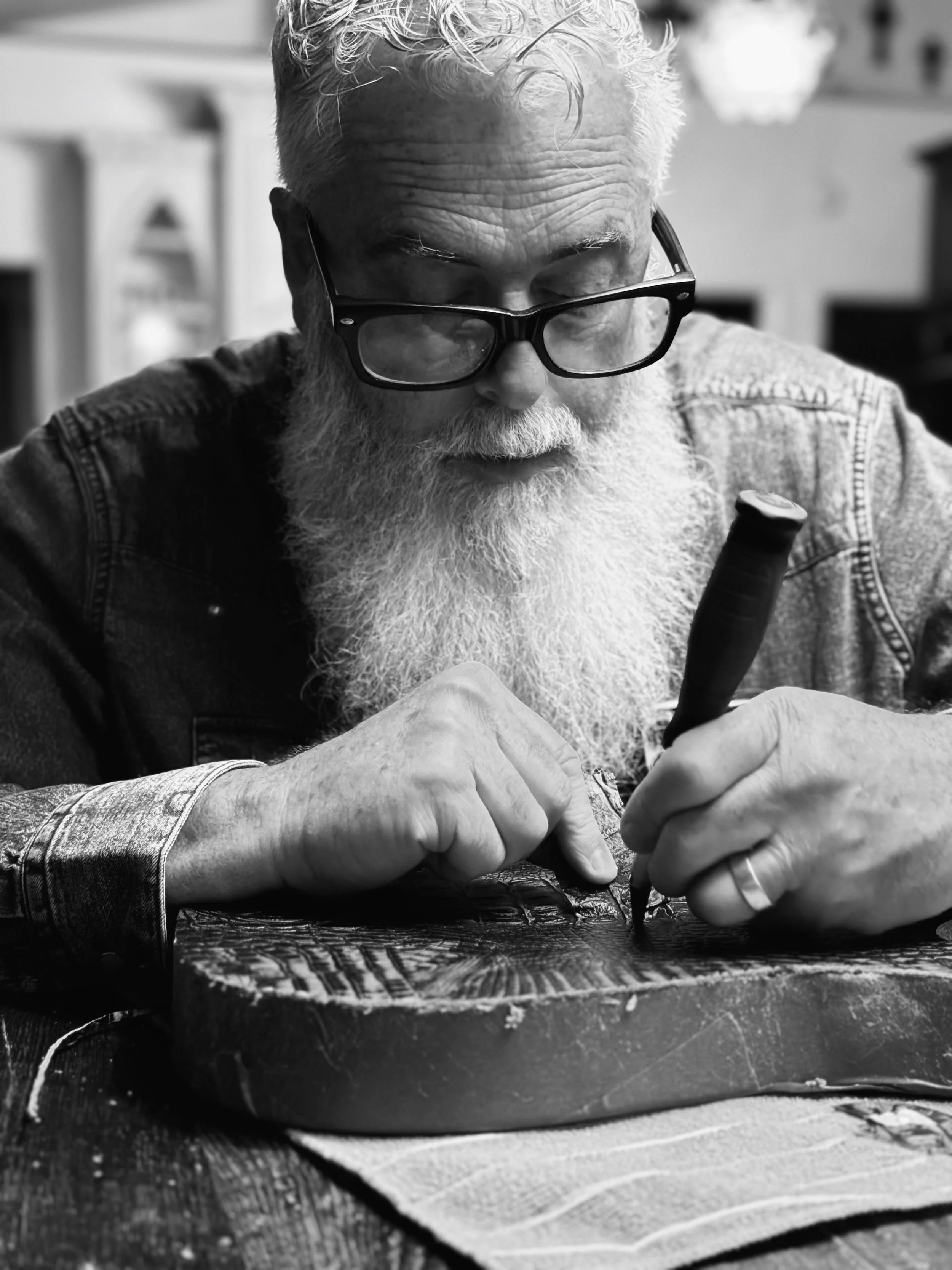 An elderly man with glasses and a white beard is carving wood with a carving tool at a table.