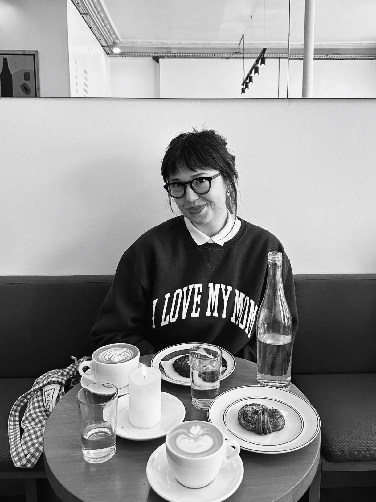 A woman with glasses, dark hair, and a checked bag sitting at a table with coffee drinks, water glasses, a bottle of water, a lit candle, and plates of pastries in a cafe with a light-colored wall and artwork in the background.
