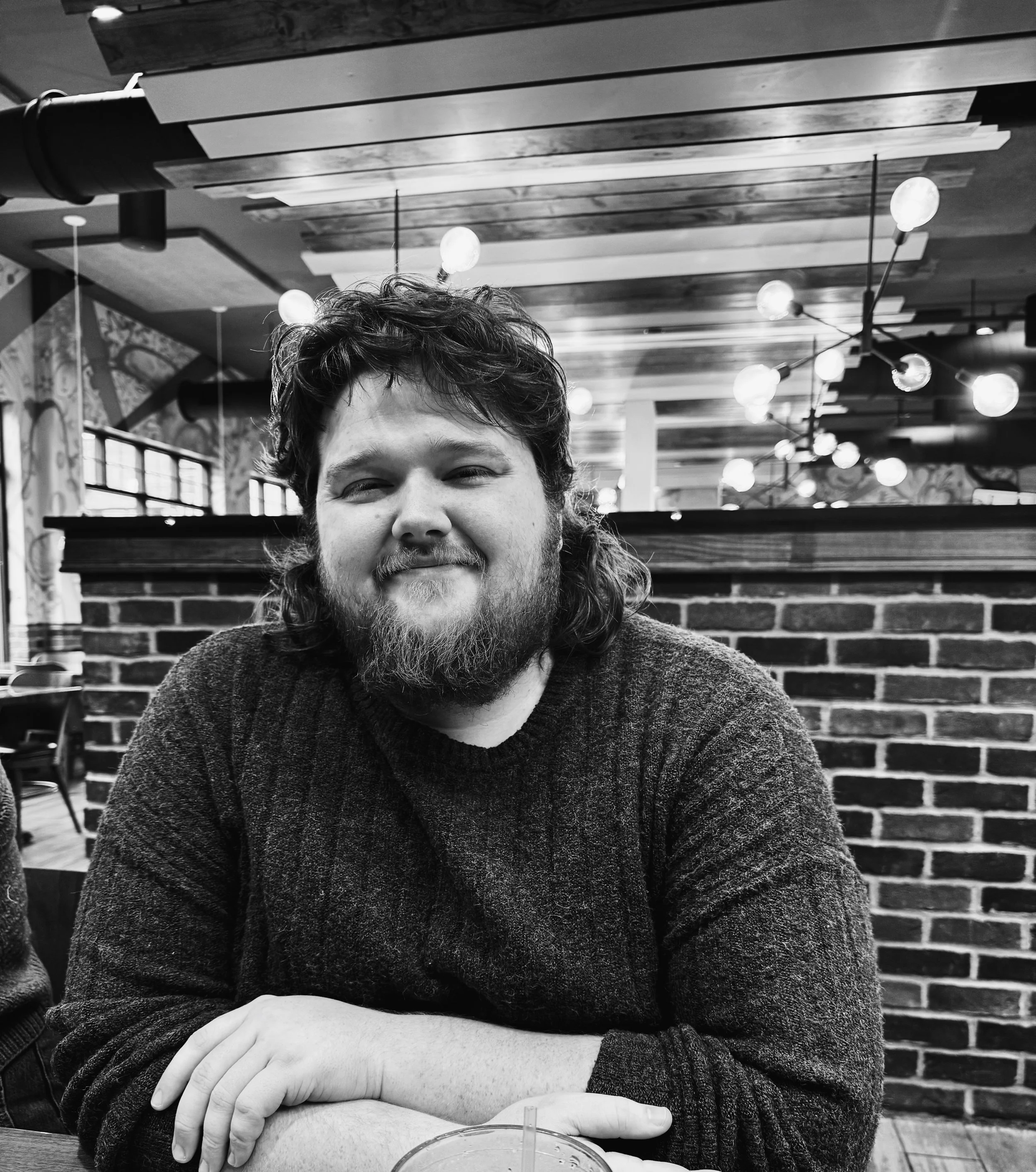 A man with a beard and curly hair smiling while sitting at a table inside a restaurant or cafe, with a brick wall behind him and decorative lighting on the ceiling.