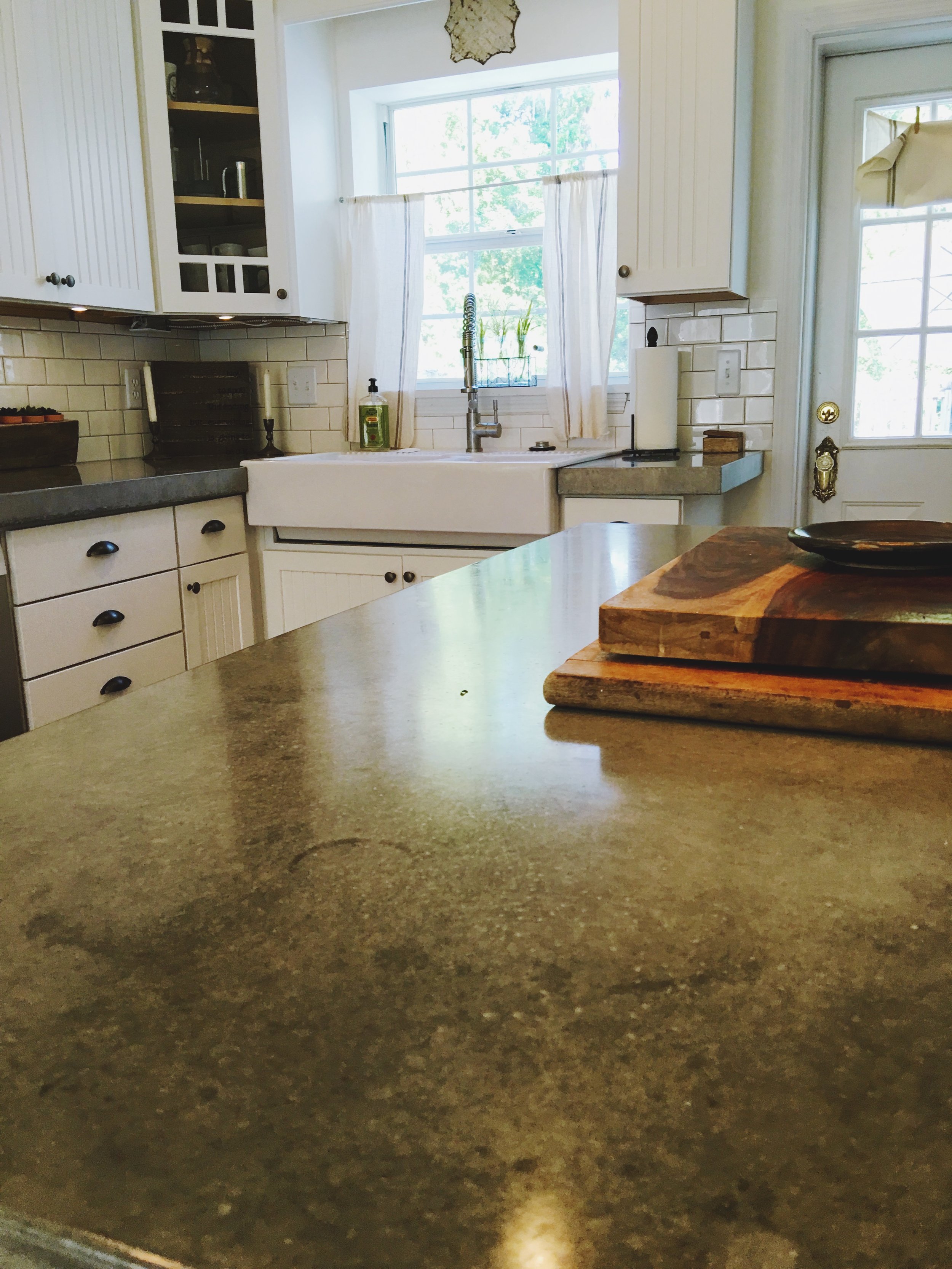 A cozy kitchen with a large window above a farmhouse sink, white cabinets, a stone countertop, and wooden cutting boards on the island.