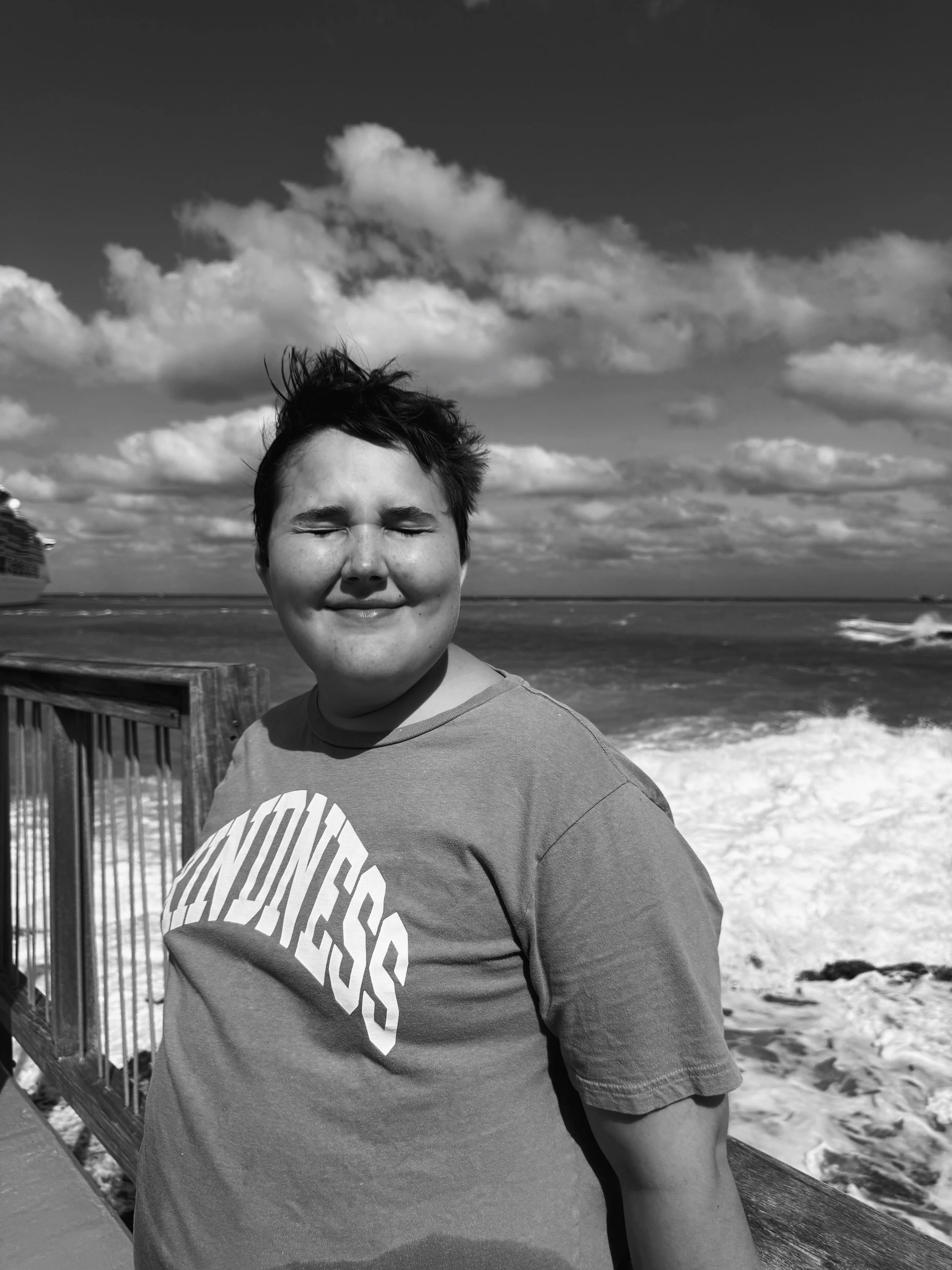 A person with short hair, eyes closed, smiling, standing on a pier near the ocean, with a cruise ship and waves in the background, under a partly cloudy sky.