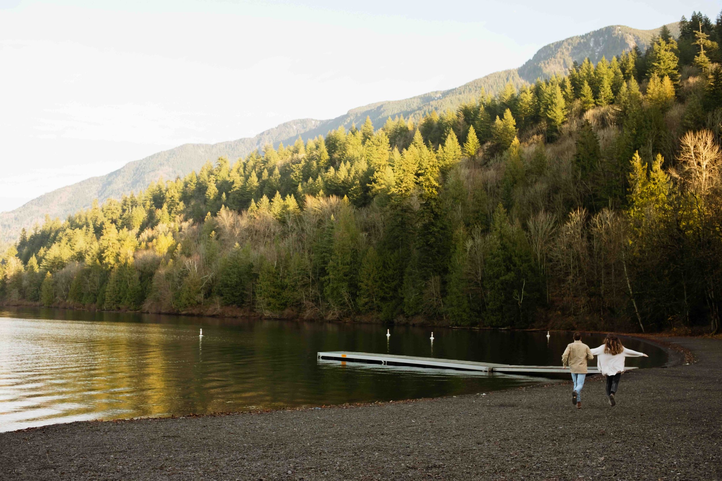 Adventurous Vancouver couple runs with arms spread wide in distance with stunning mountain lake background