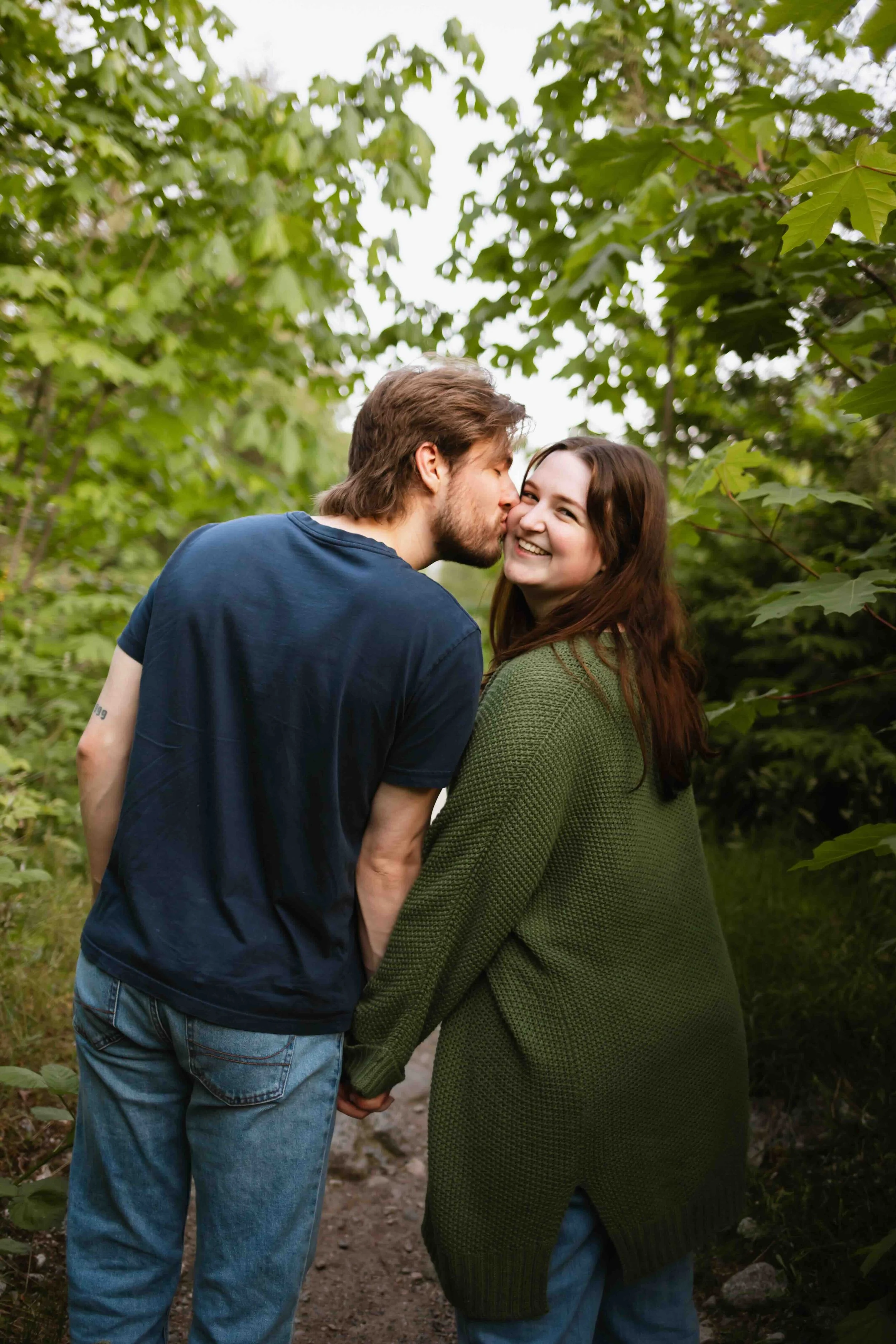 Vancouver woman smiles over her shoulder, holding hands with her fiance who kisses her cheak