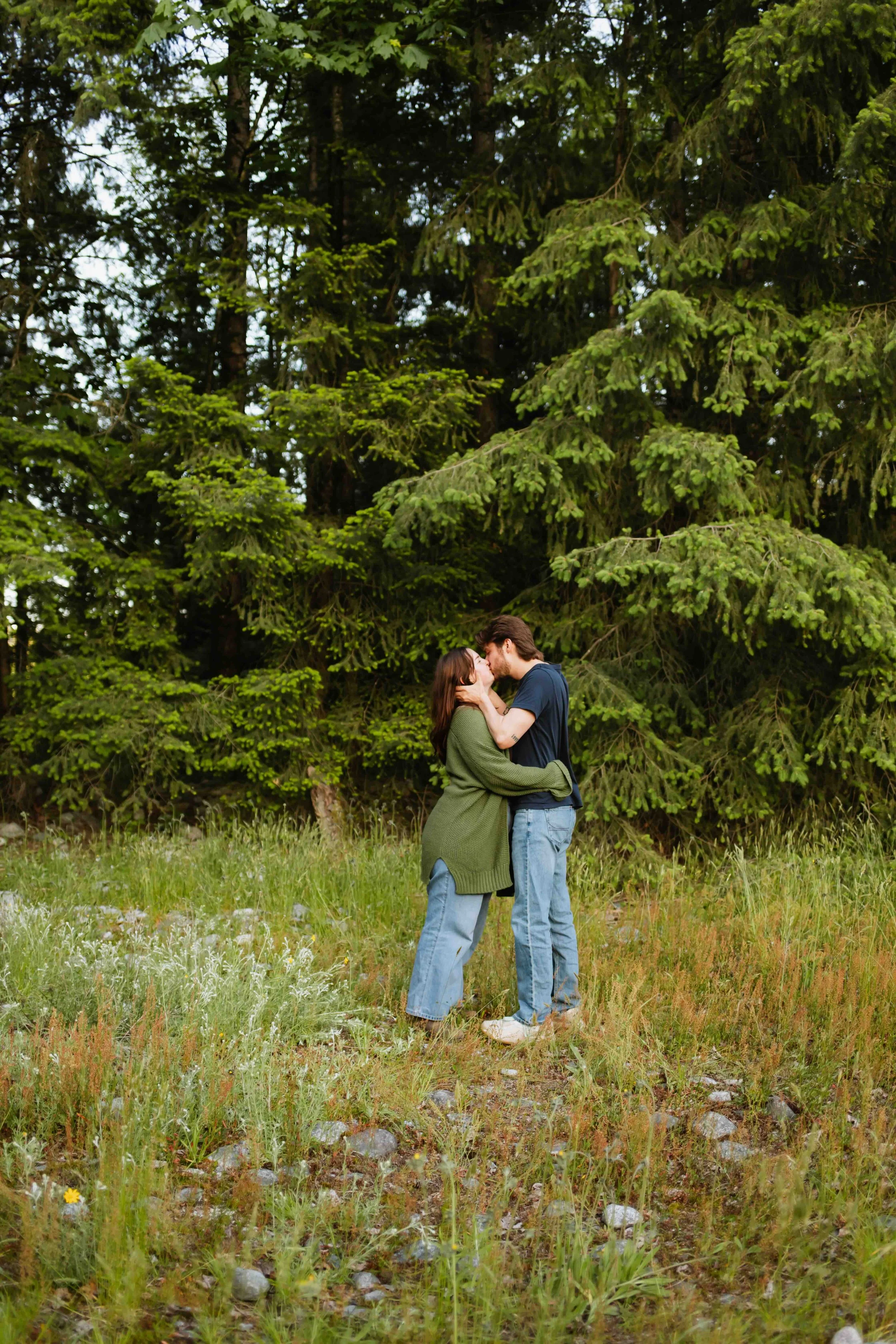 Vancouver couple kisses in rugged field with a lush forest behind them