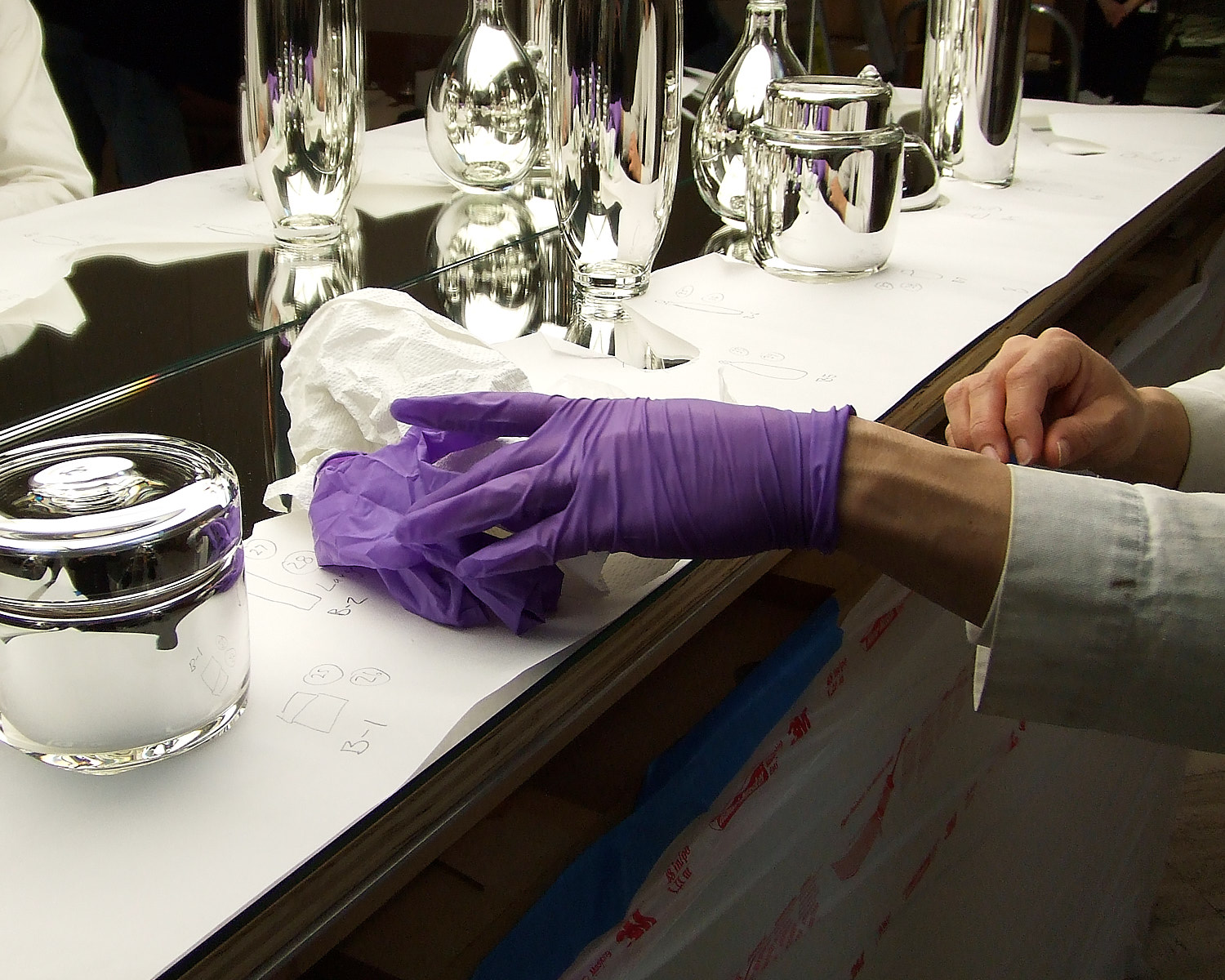 Person wearing a purple glove cleaning a reflective surface with a cloth beside polished metal objects on a paper-covered table.