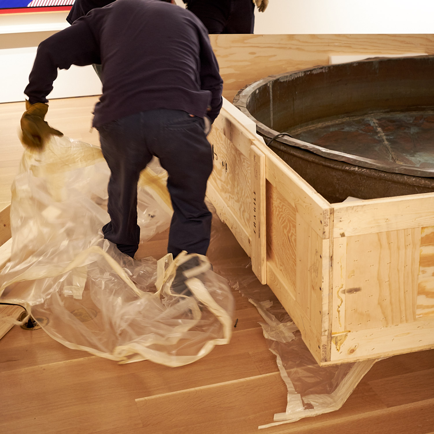 Person unpacking a large circular object from a wooden crate, removing plastic wrapping, indoors.