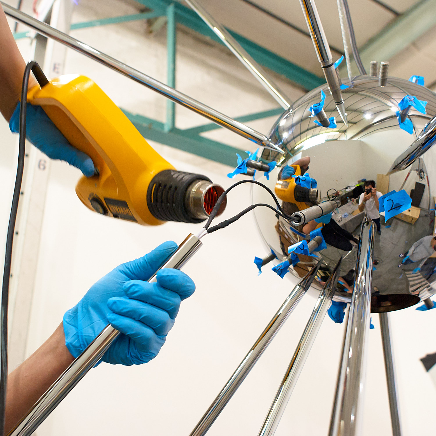 Person using a heat gun on a mirror-like metallic sphere with blue tape, wearing blue gloves.