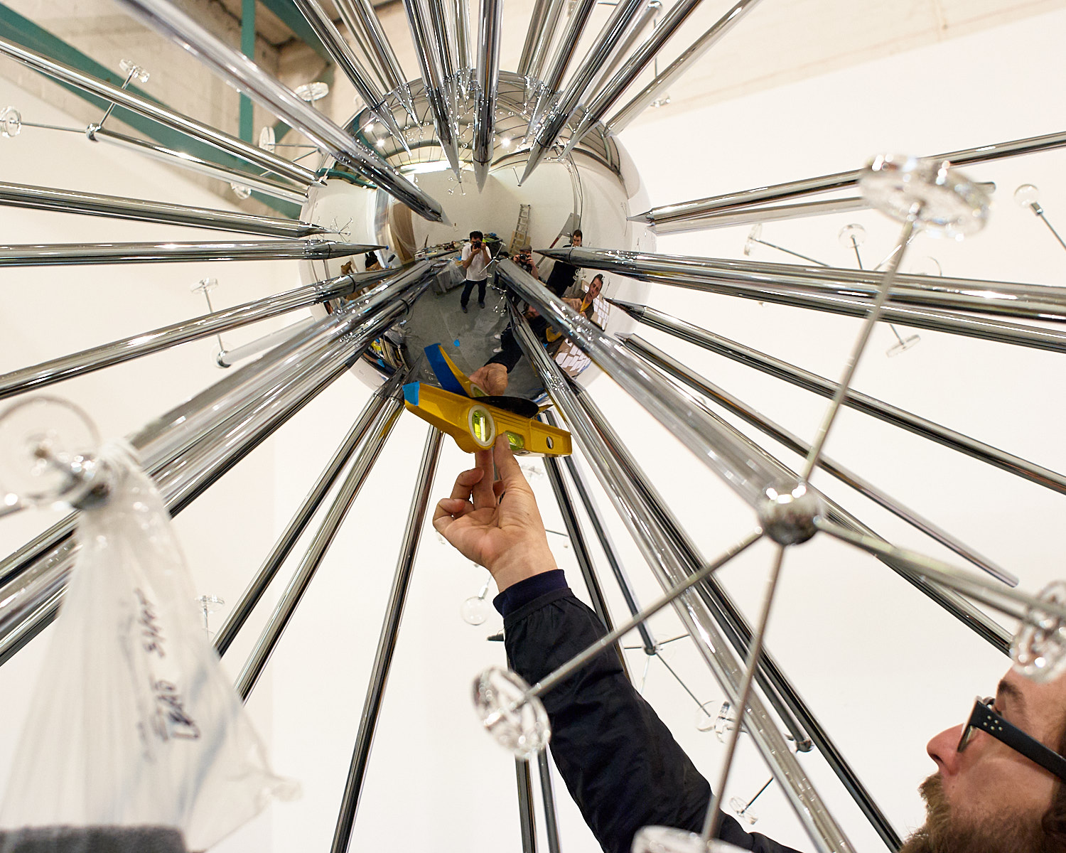 A person using a level tool to adjust a large, metallic, spherical sculpture with multiple rods extending outward.